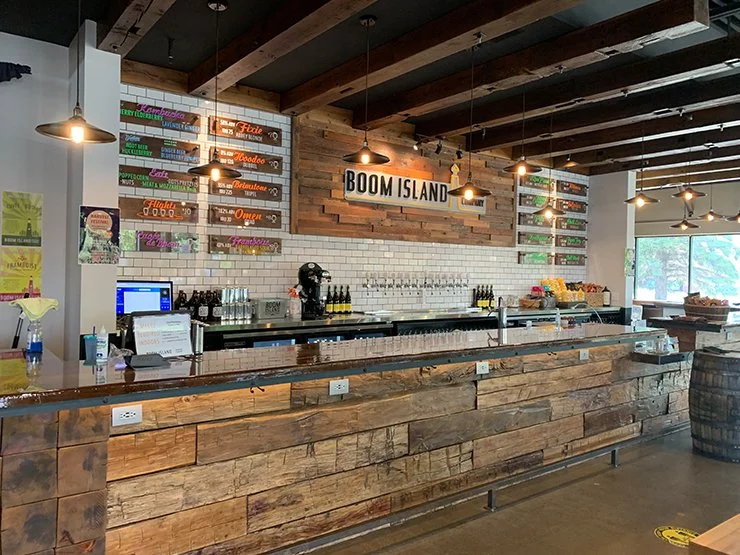 Rustic brewery taproom interior with wooden bar, beer taps, hanging pendant lights, and a large "ROOM ISLAND" sign.