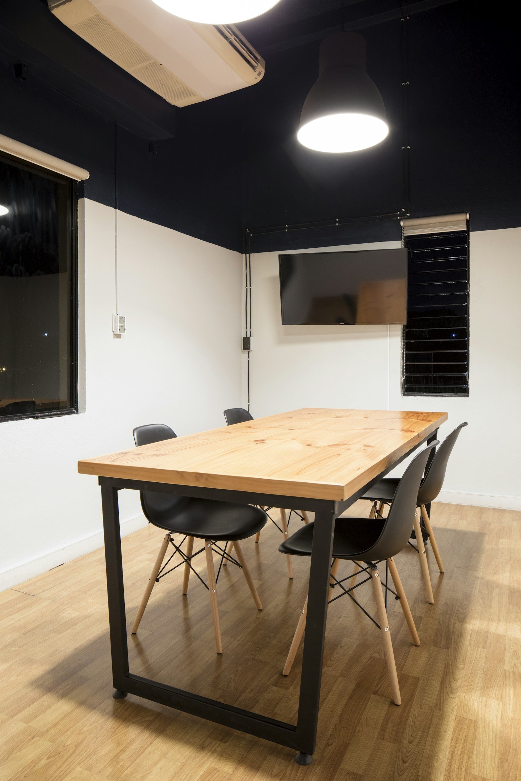 A meeting room with a wooden table, six black chairs with wooden legs, a wall-mounted TV, black blinds, a window, and a large ceiling lamp in a modern design.