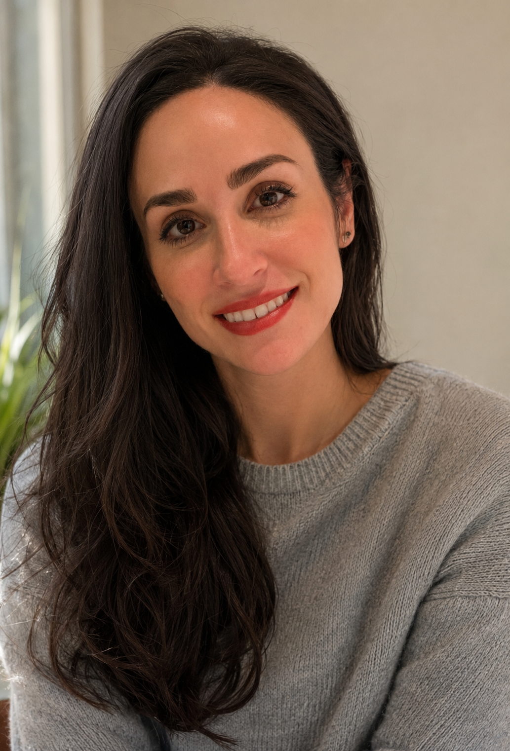 A woman with long dark hair wearing a gray sweater, smiling at the camera inside a room with a plant and light-colored wall in the background.