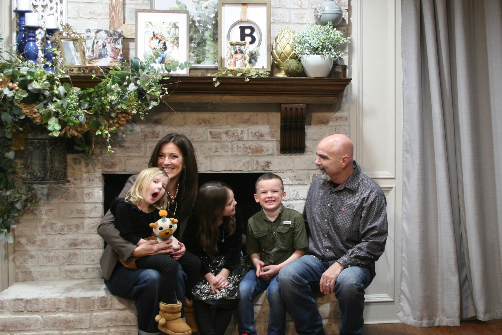 A group of five people, including two adult men and women and three children, are sitting on a brick hearth in front of a white brick fireplace. The woman on the left is holding a plush cow toy and is smiling. The children are sitting next to the women, with the girl on the left wearing black and leopard print shoes, and the boy in the middle wearing a green shirt. The man on the right is smiling and looking at the children. The background features a mantel decorated with framed photos, plants, and decorative objects, with a brick wall and a beige curtain on the right side.