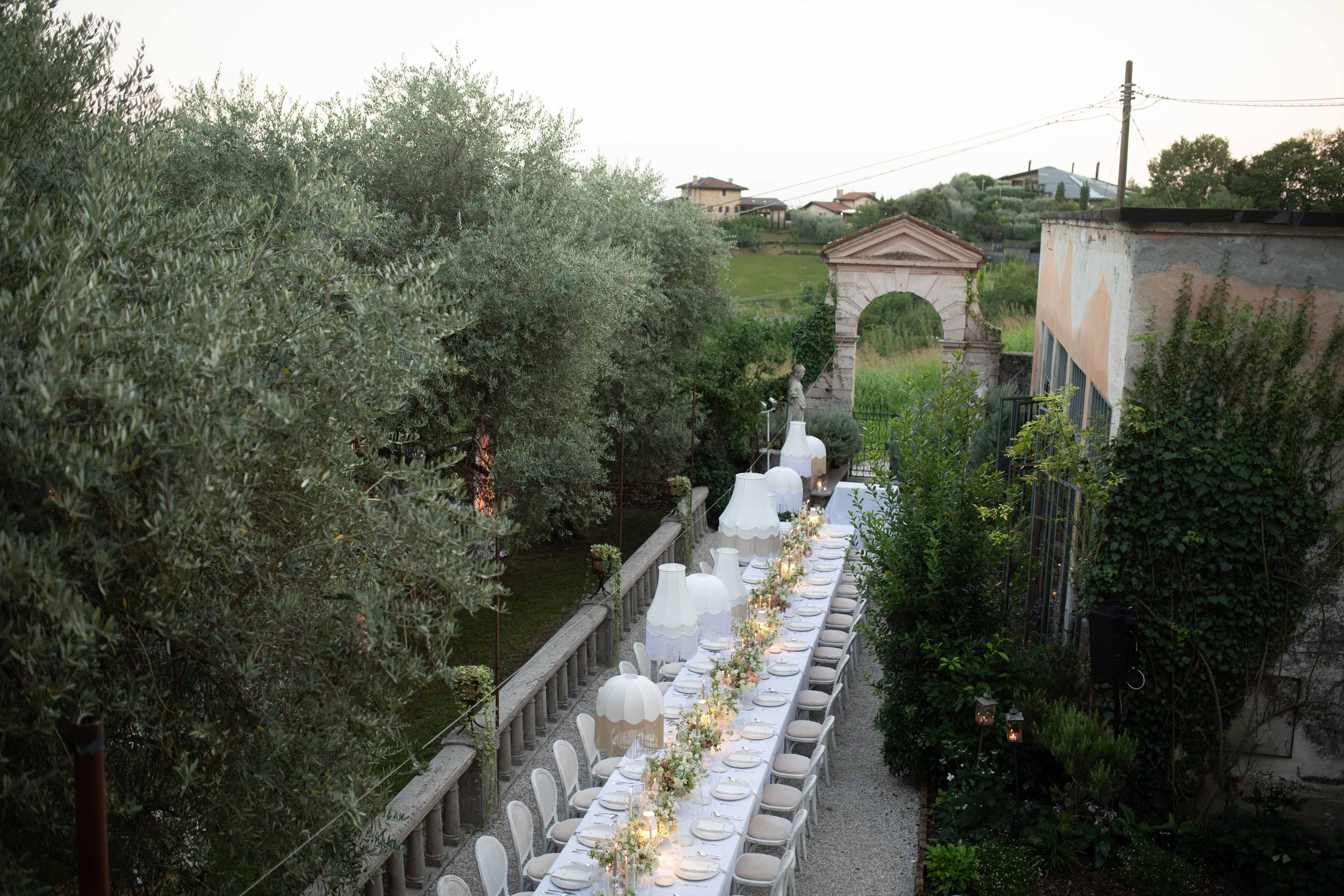 Outdoor party setup with a long table decorated with white tablecloths, floral centerpieces, and large white lamps, surrounded by green trees and a stone archway in a lush garden setting.