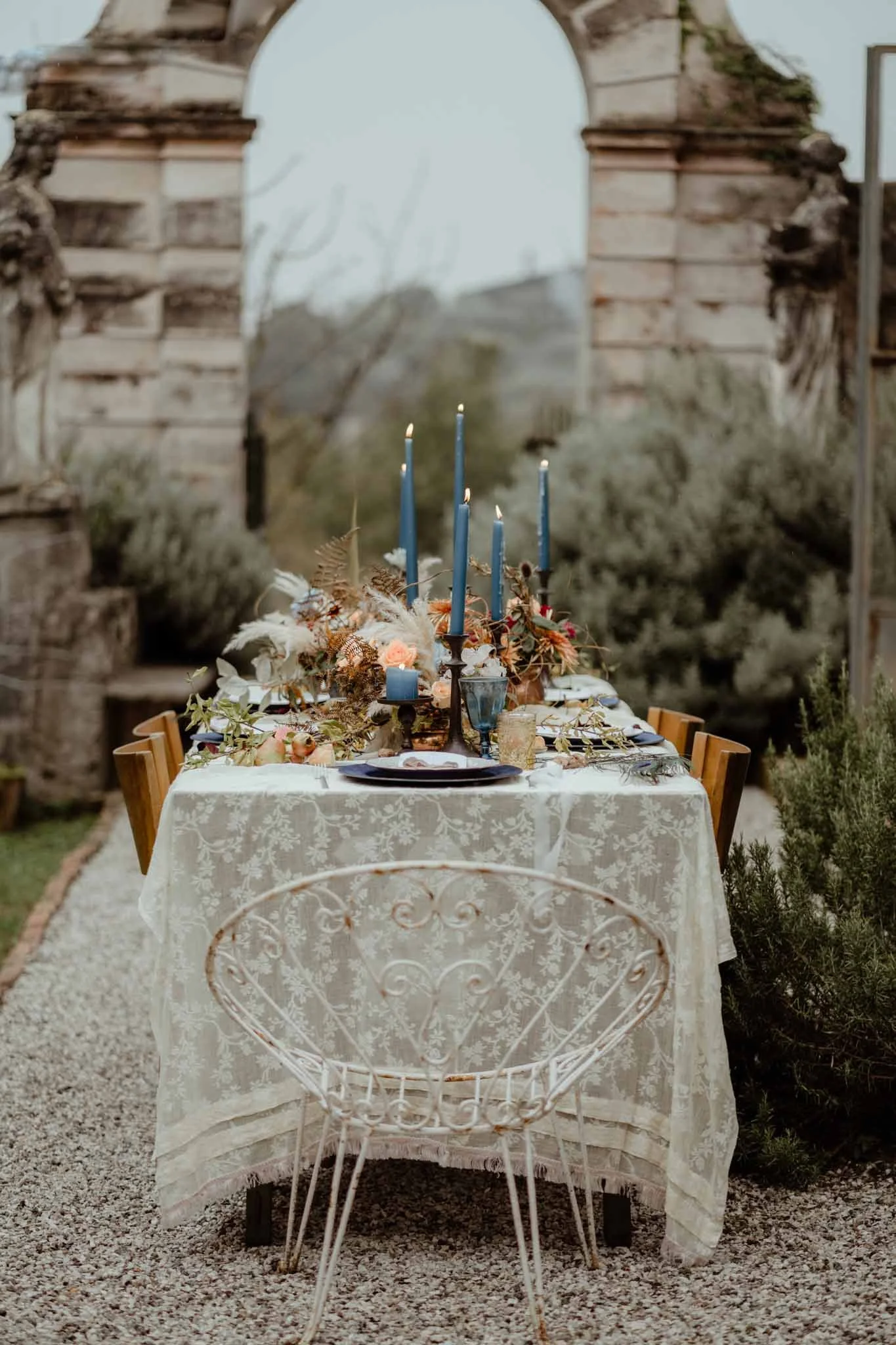 Outdoor dining table with a white lace tablecloth, surrounded by wooden chairs, decorated with flowers, candles, and tableware, set in a garden with a stone archway in the background.