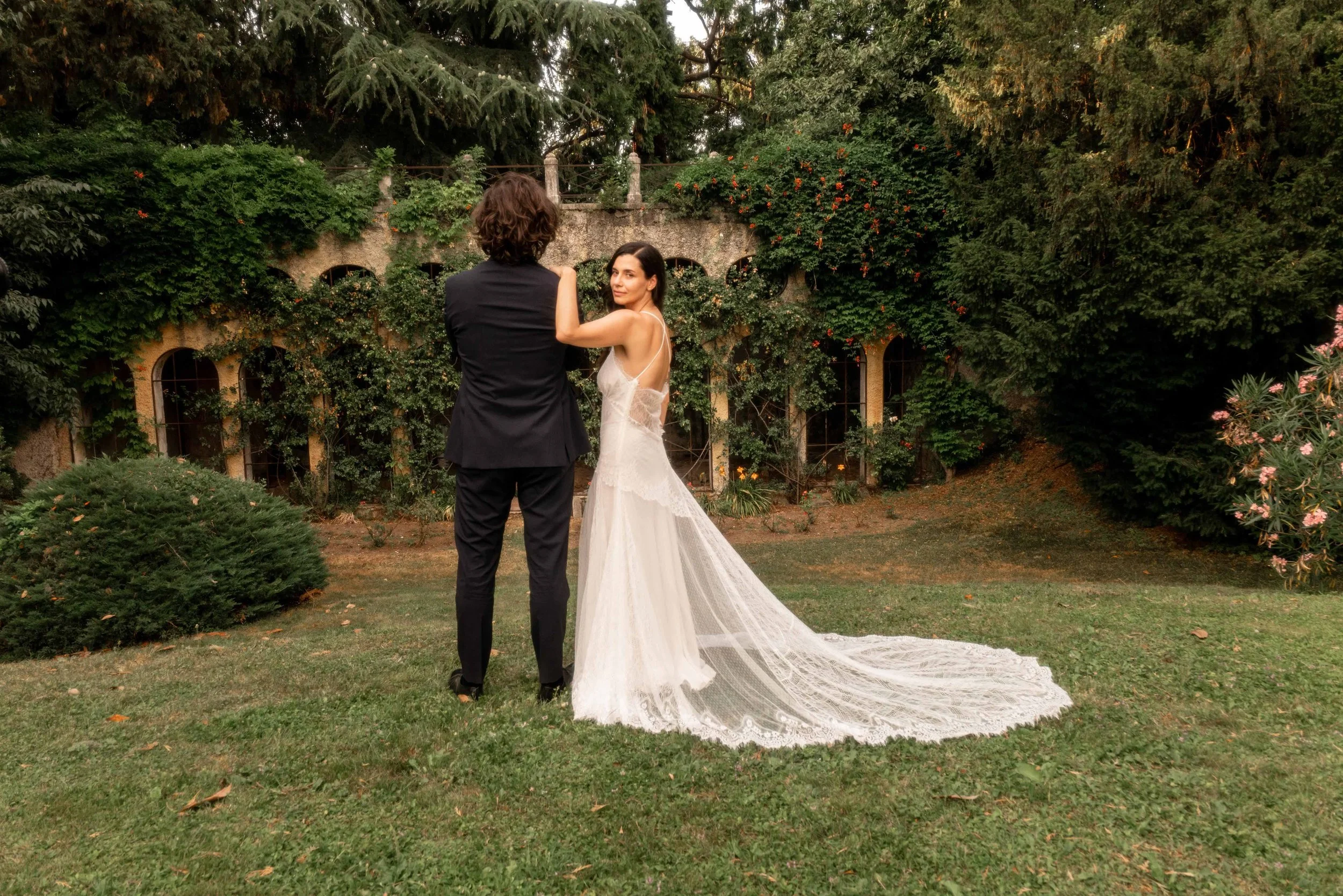 A bride and groom holding hands and facing each other in a lush garden with greenery and flowering plants, with the bride wearing a white wedding dress and the groom in a black suit.