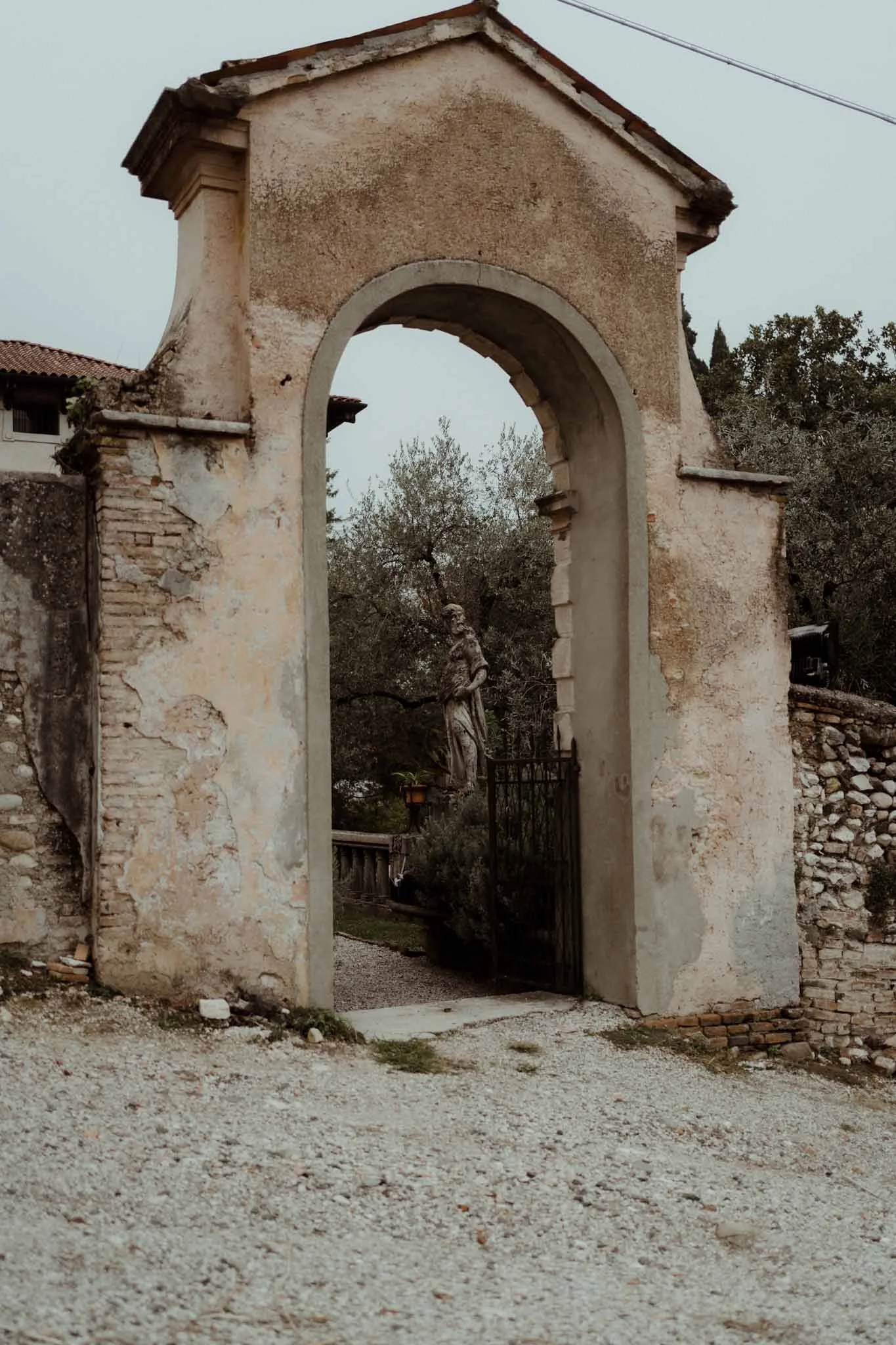 Old stone and plaster archway entrance to a historic site, with trees and a pathway visible through the arch.