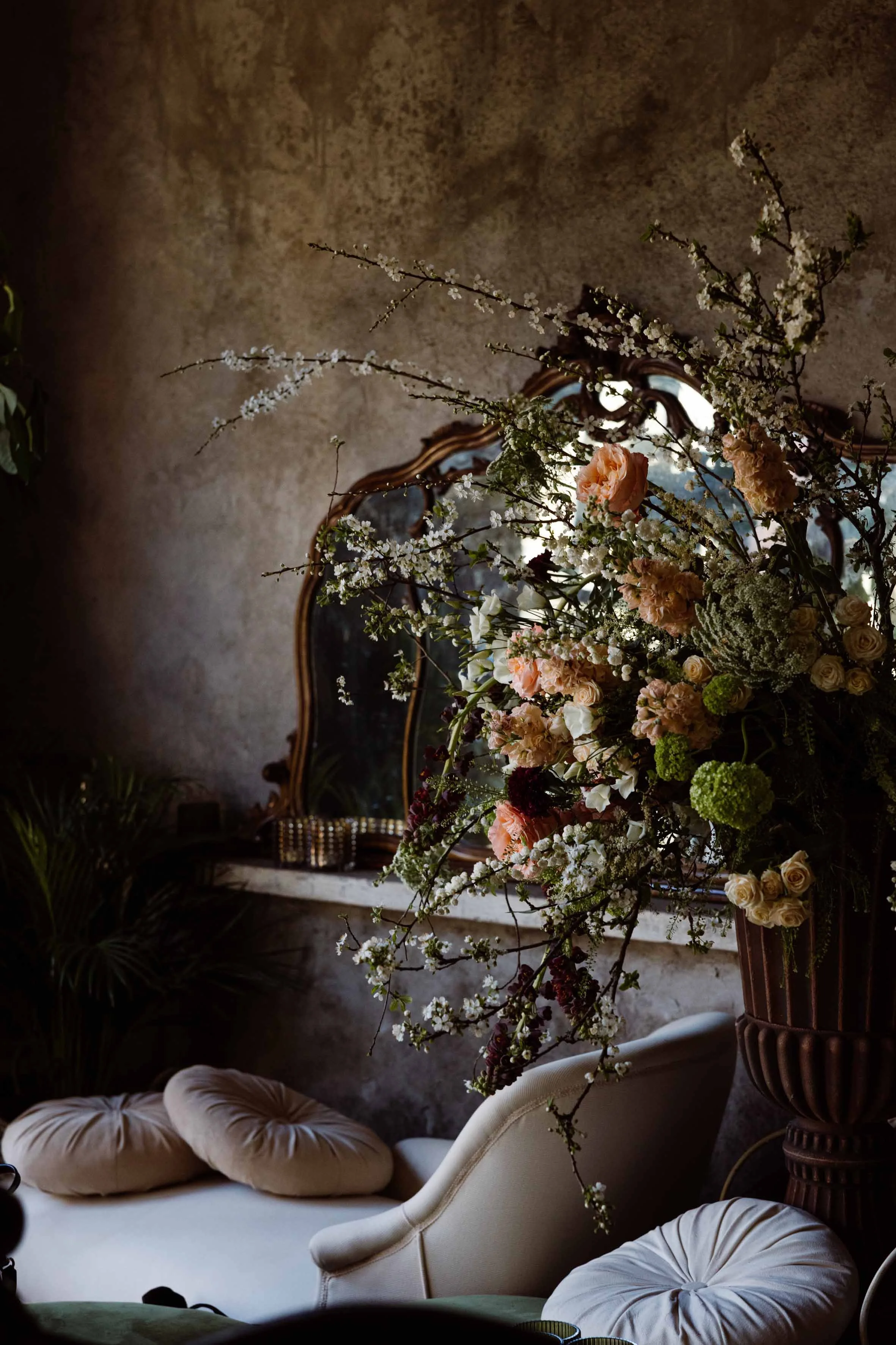 A floral arrangement with pink and white flowers on a table in front of a vintage mirror in a dimly lit room.