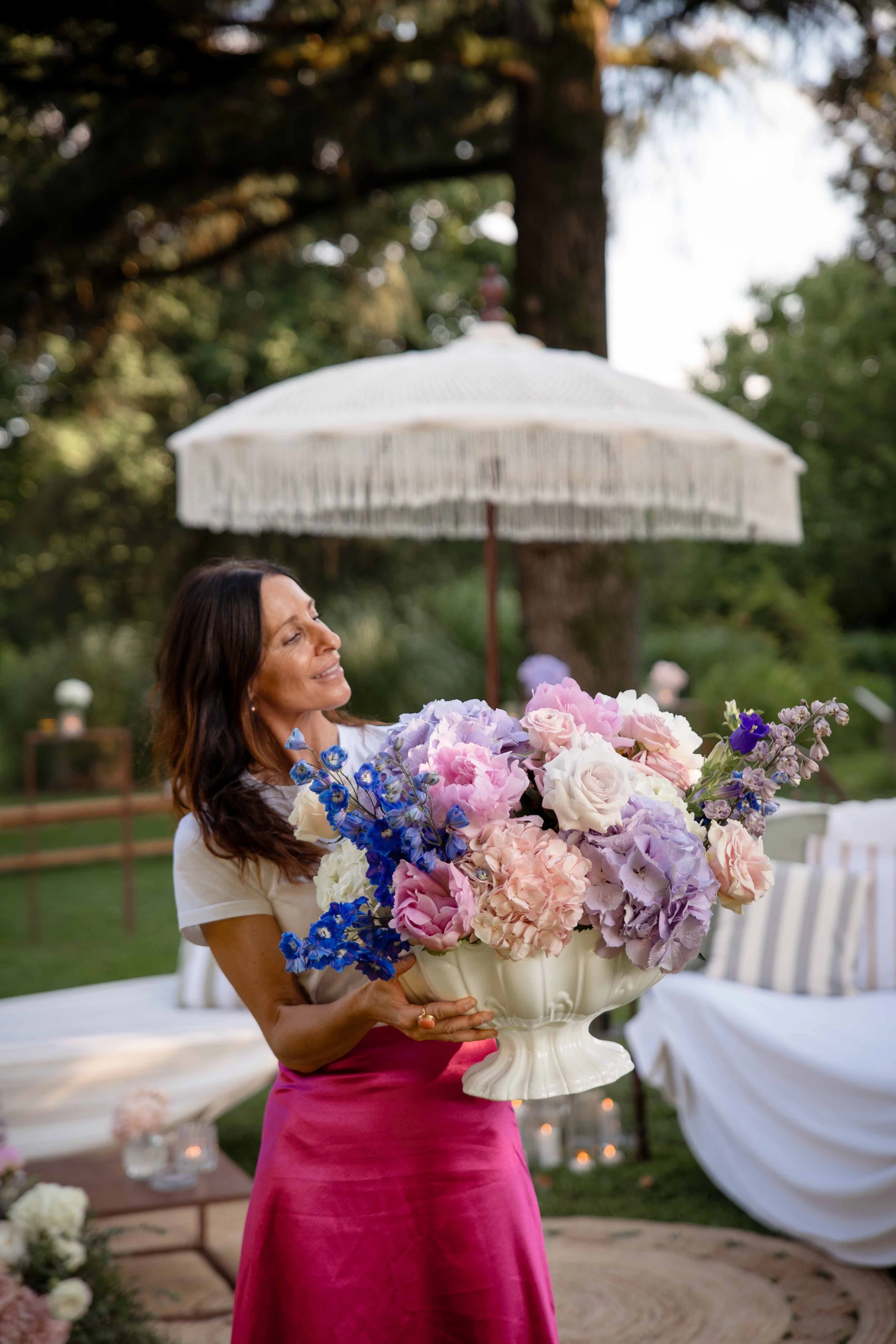 A woman in a white top and pink skirt holding a large white vase of pink, white, purple, and blue flowers at an outdoor event with umbrella, trees, white chairs, and lit candles in the background.