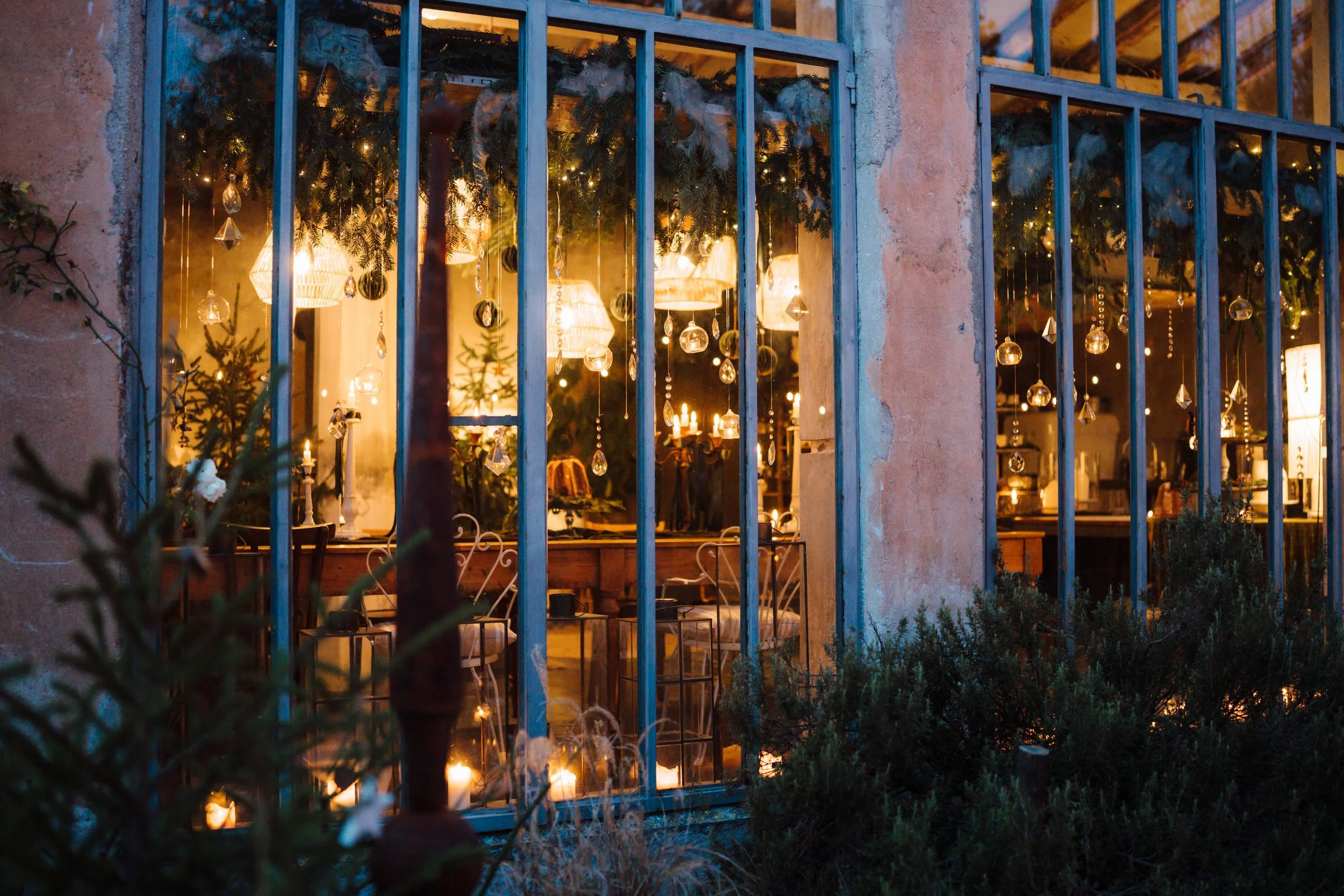 View through a window with metal bars, showing cozy indoor lighting and Christmas decorations inside, illuminated with warm lights, with greenery visible outside.