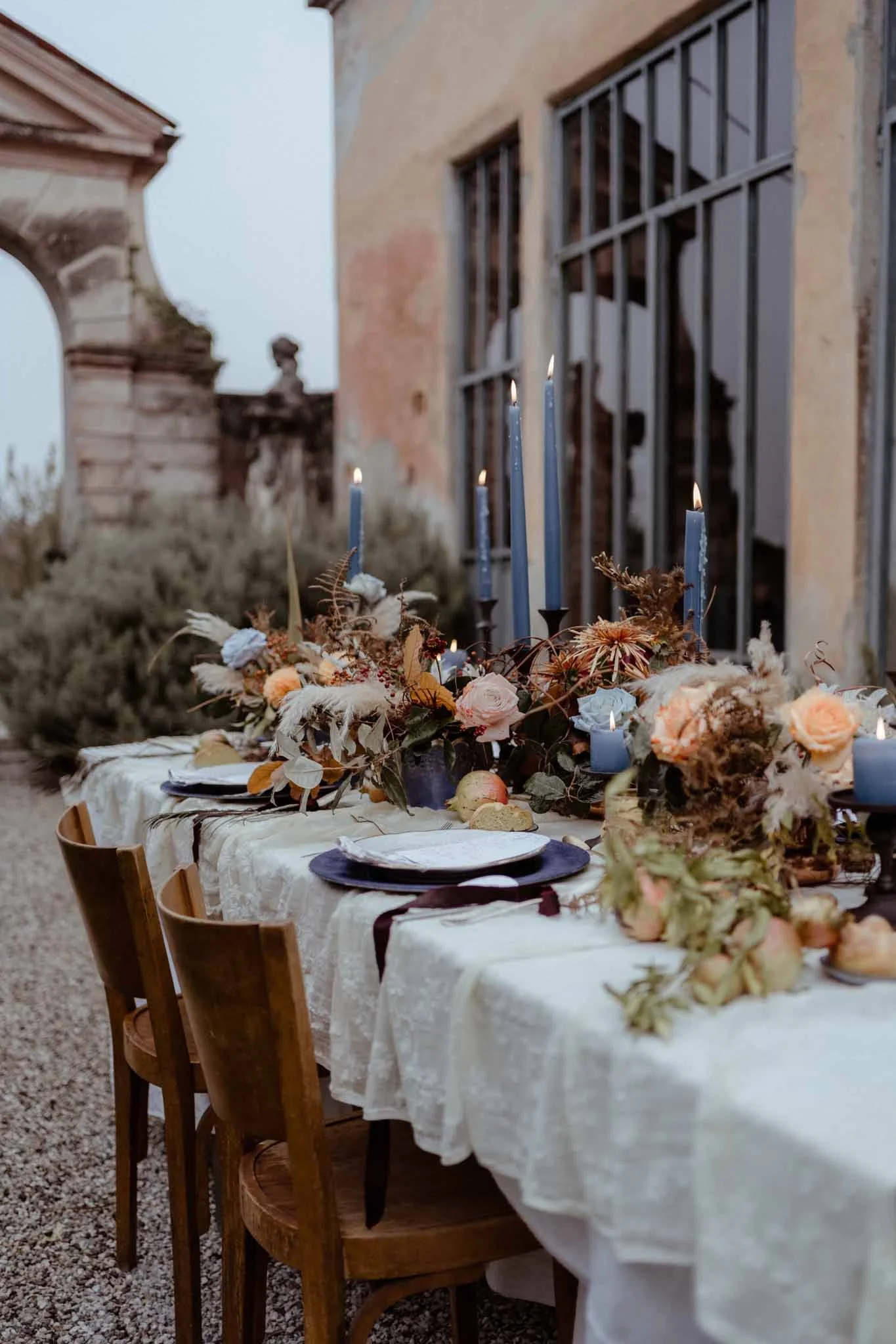 An outdoor dining table decorated with candles, flowers, and tableware, set against an aged building with barred windows, in a rustic, historic setting.