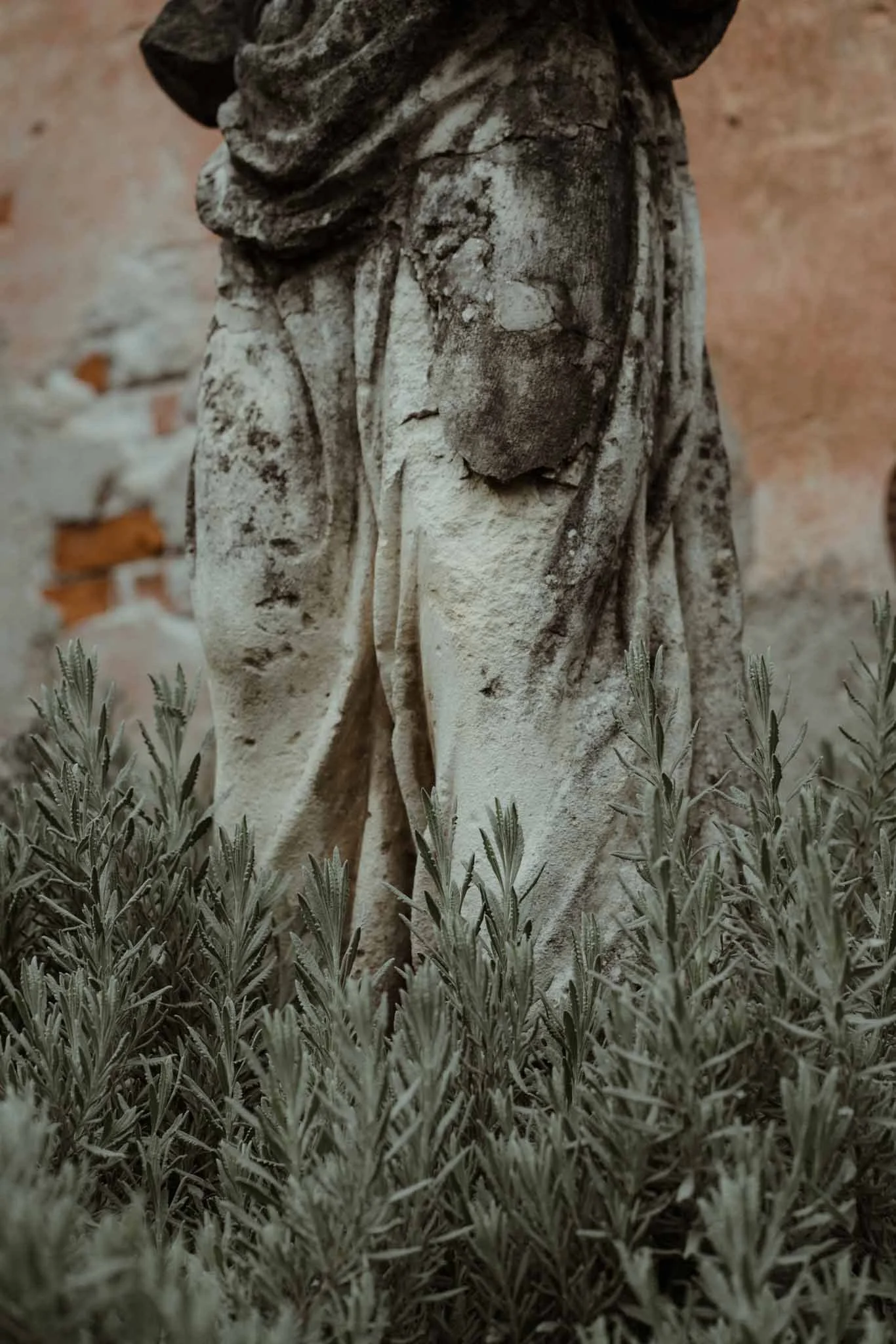 Close-up of a weathered stone statue surrounded by green plants, with a brick wall in the background.