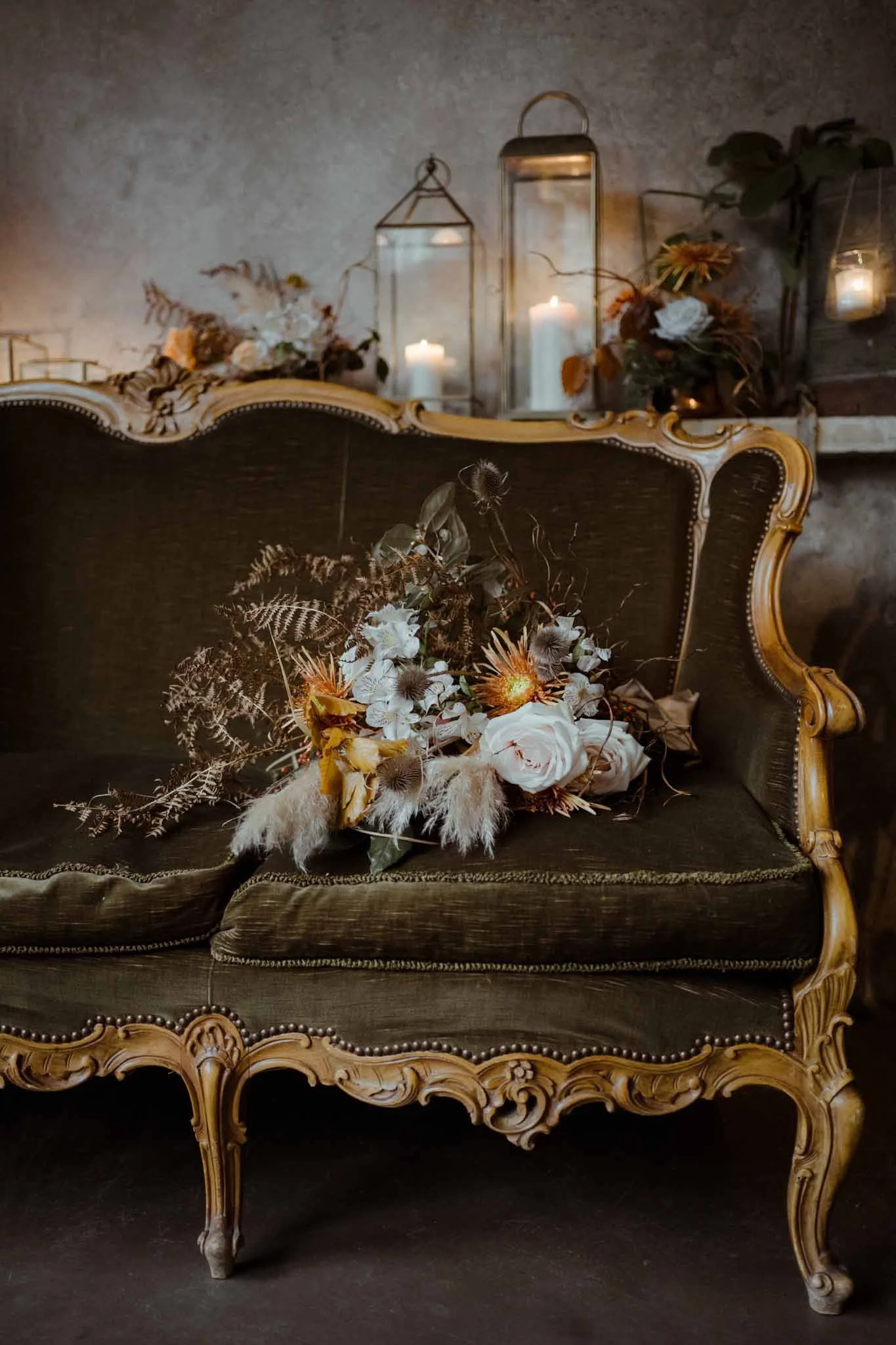 Vintage-style dark velvet sofa with ornate wooden frame with gold accents, on a dark floor. Behind the sofa, a shelf with lit candles in glass lanterns, dried flowers, and leaves is displayed against a gray wall, creating a cozy, rustic ambiance.