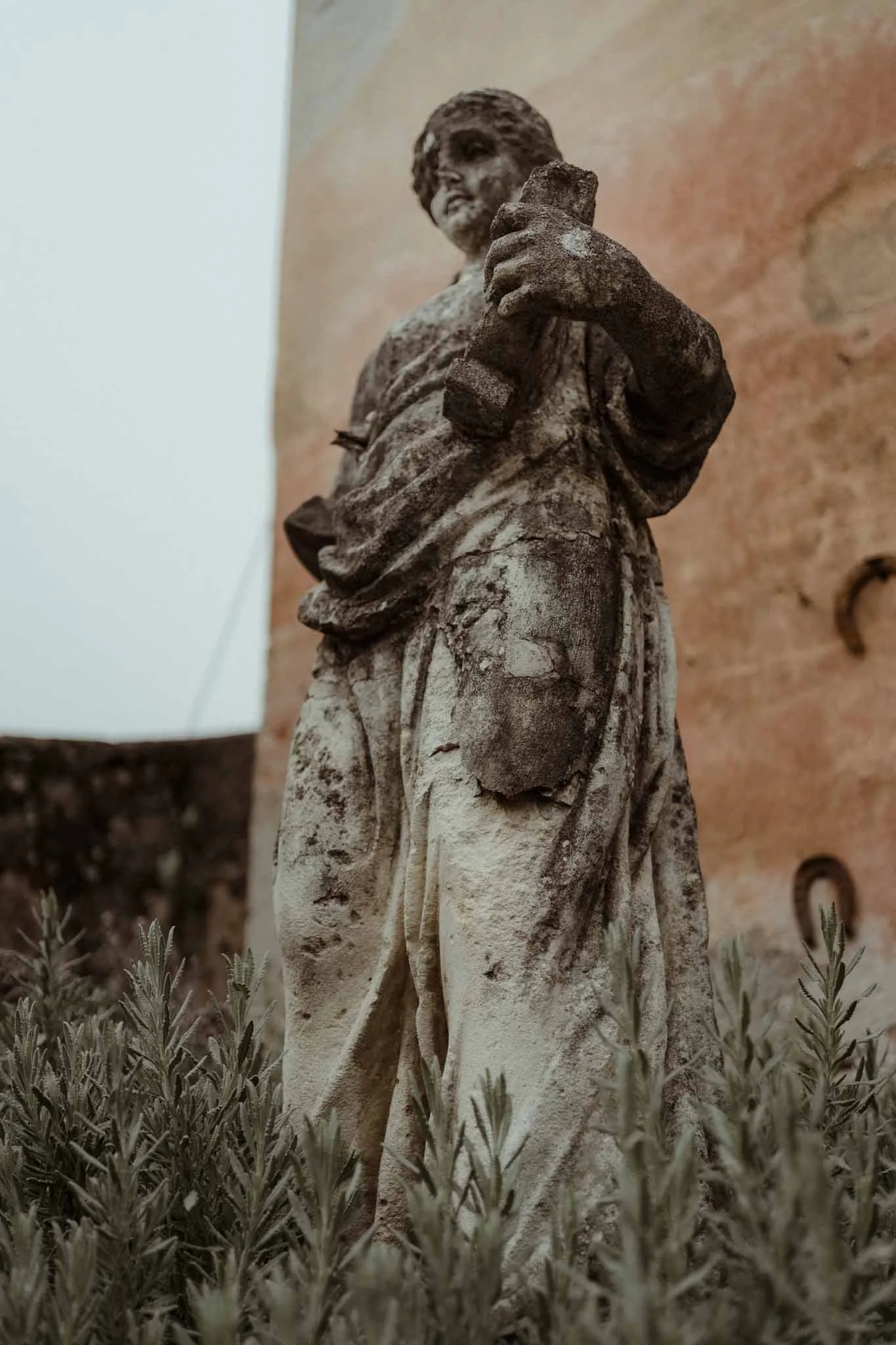 A weathered stone statue of a person with short hair, holding an object, standing among bushes.