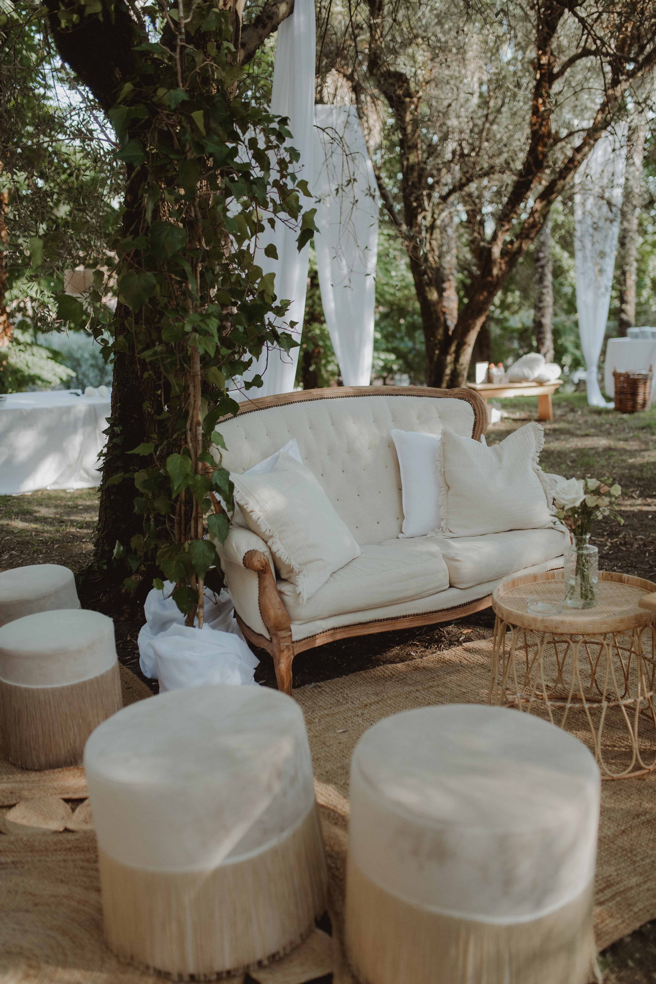 Outdoor seating area with white cushioned vintage sofa, small round wooden table, and cylindrical wooden stools on a woven rug, surrounded by trees and draped white fabric, creating a cozy, natural setting.