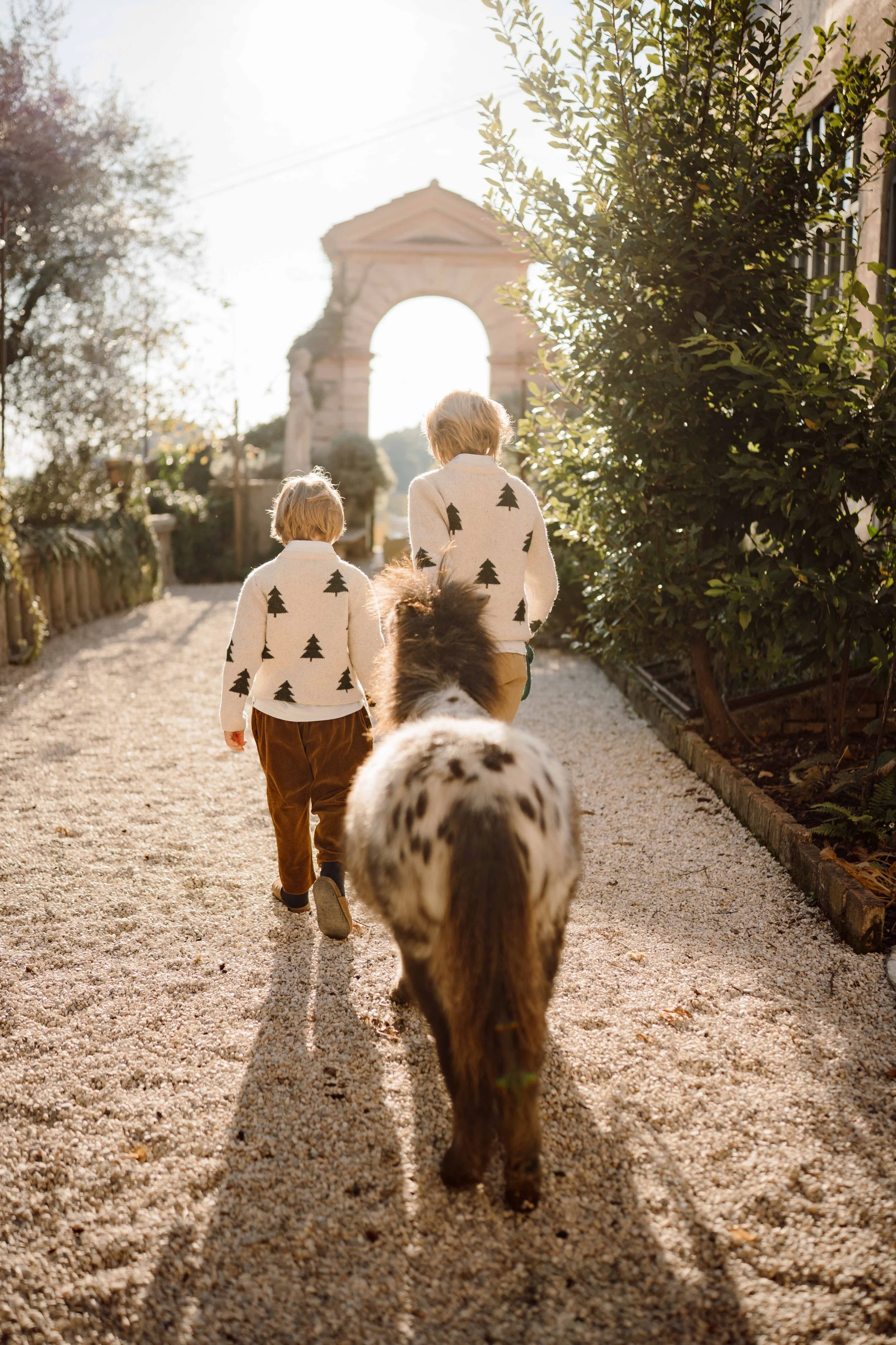 Two children walking down a gravel path with dogs, wearing matching sweaters with tree patterns. The scene is illuminated by warm sunlight with trees and an archway in the background.