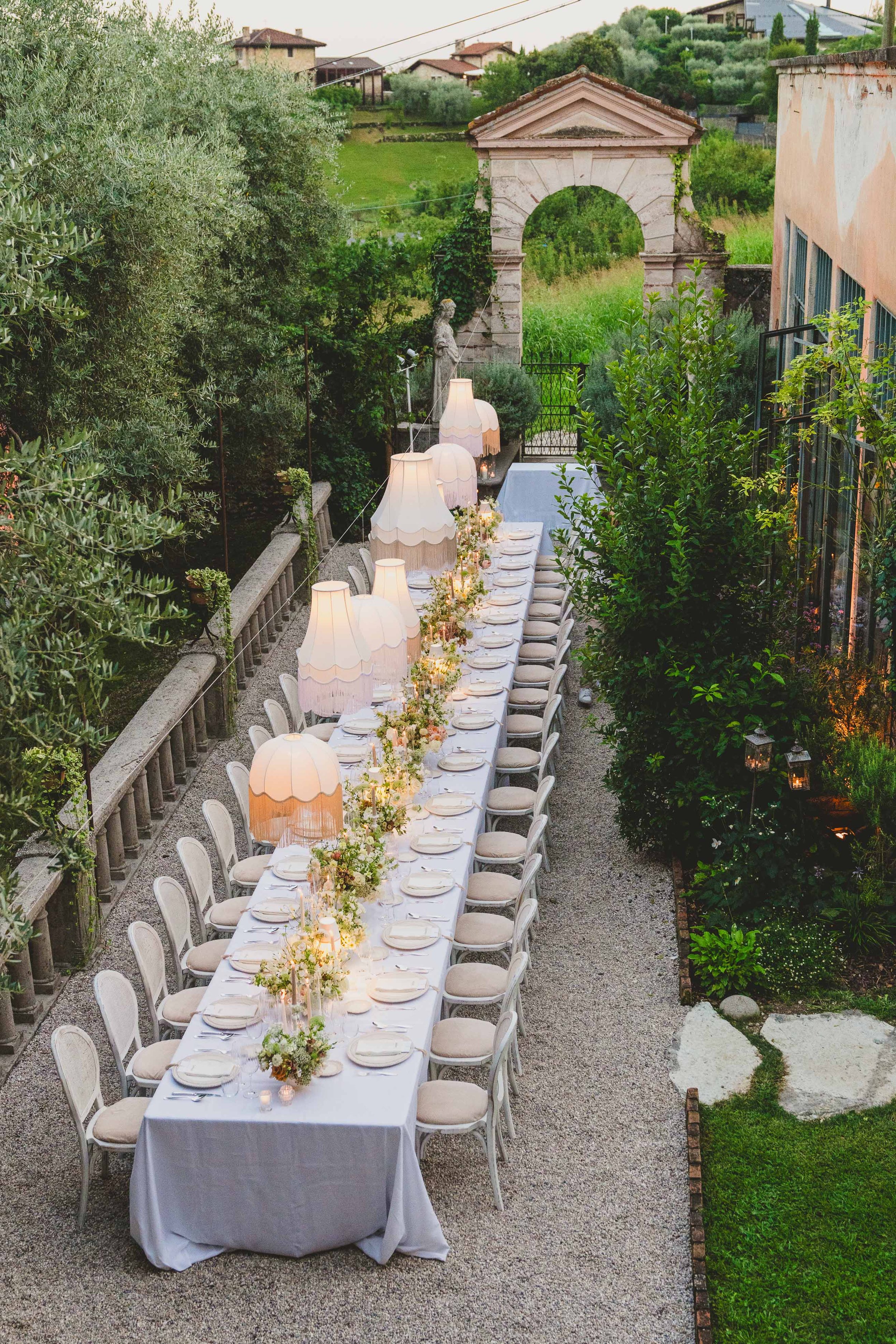 Long dining table set outdoors with white tablecloth, flowers, and vintage lamps, surrounded by greenery in a garden with an archway in the background.
