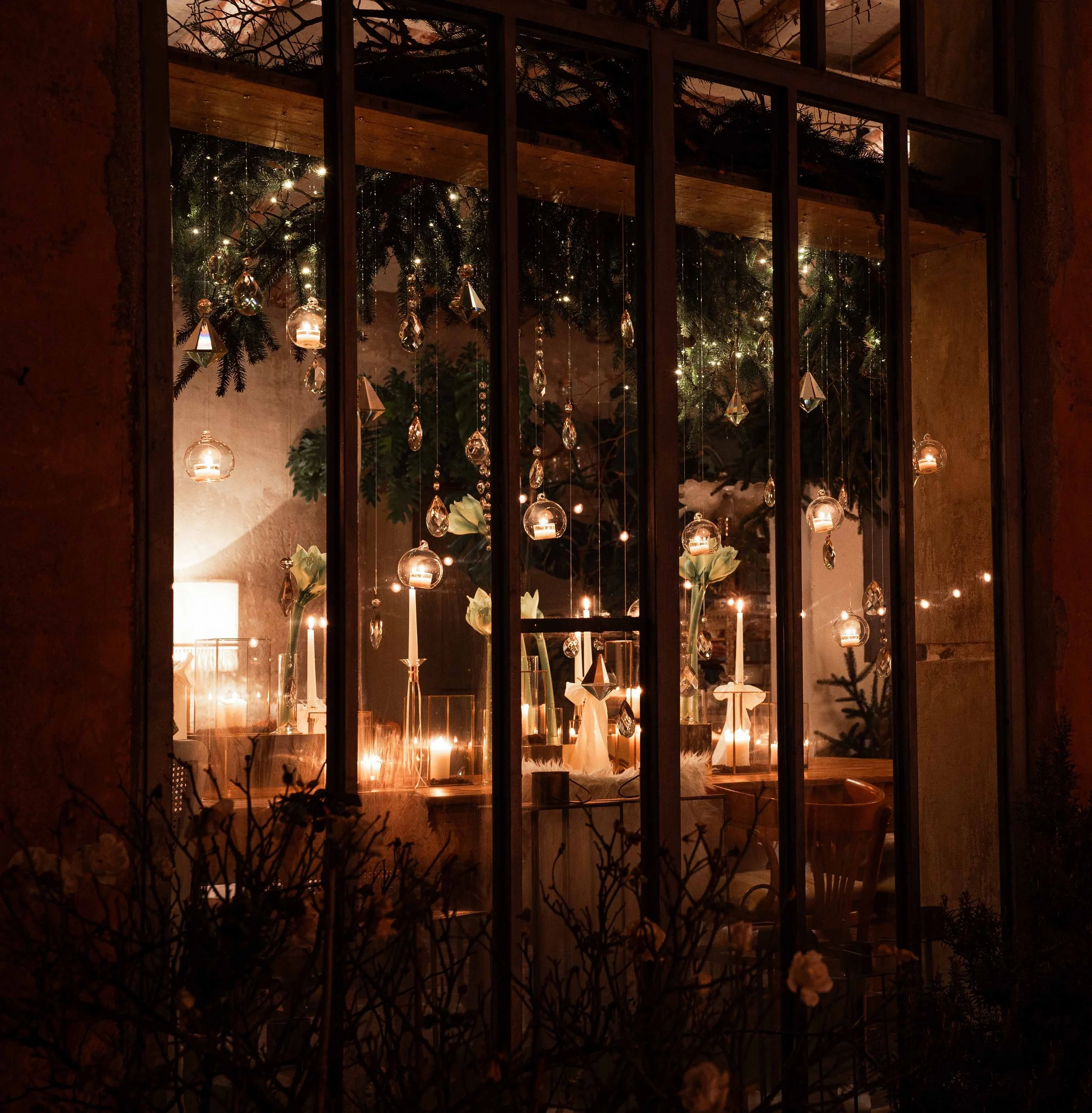 A window with decorative hanging crystals and candles inside, viewed through some foliage at night.