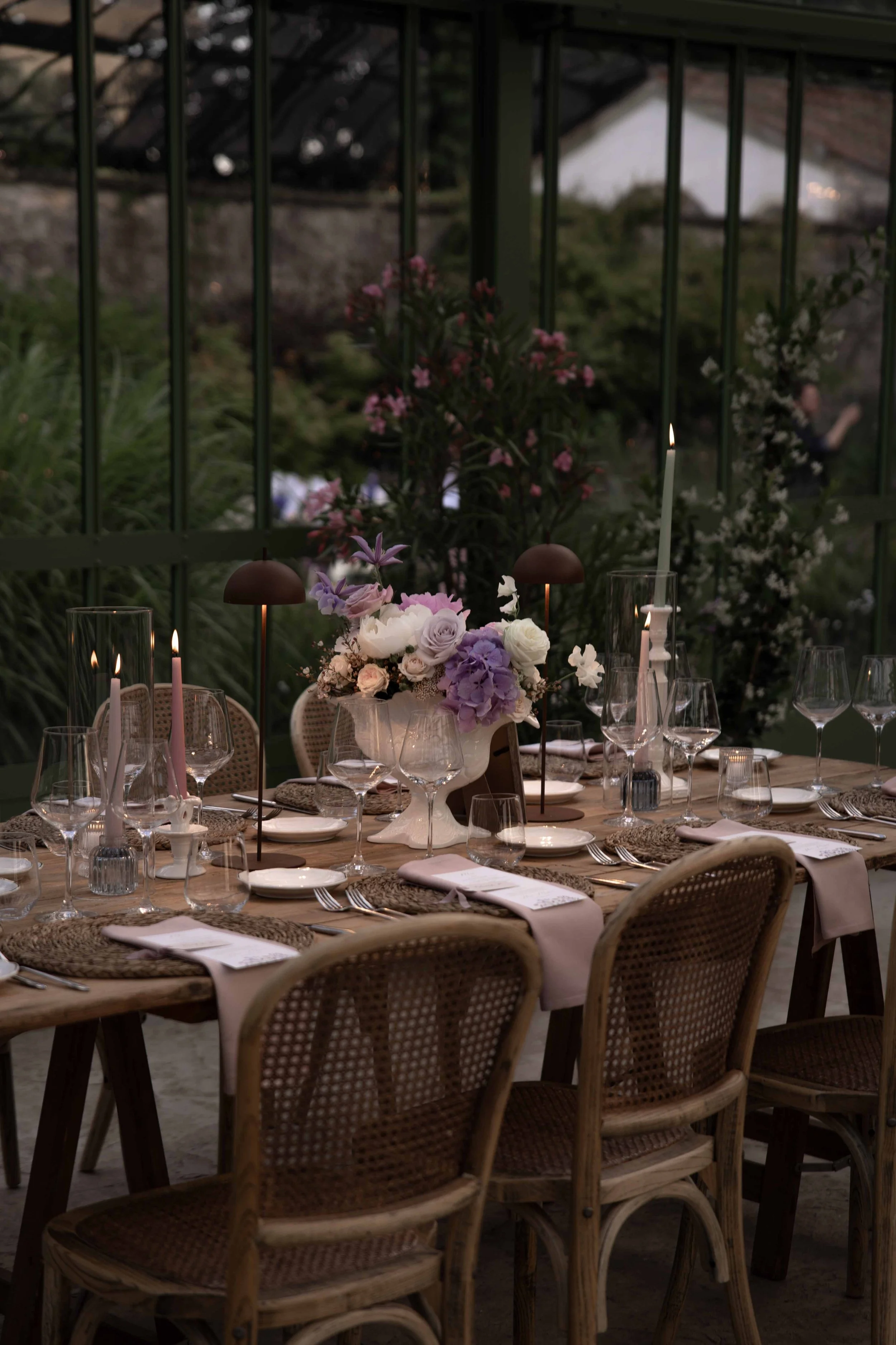 A dinner table set with elegant plates, glasses, candles, and floral arrangements, situated outdoors with a railing and greenery in the background.