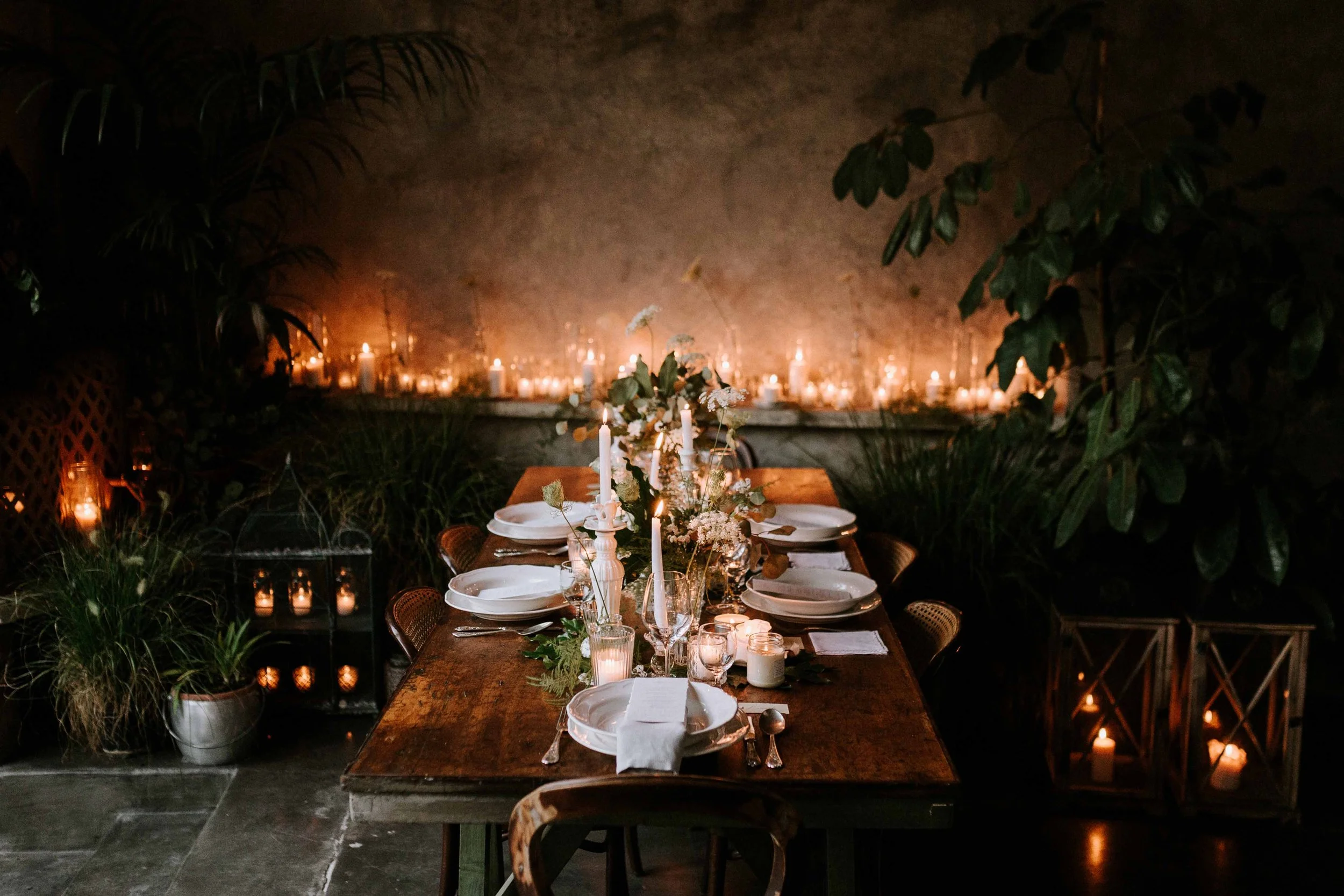 A dimly lit indoor dinner setting with a long wooden table, white plates, glasses, and silverware. The table is decorated with candles and greenery. The background features numerous candles and lush green plants, creating a cozy and intimate atmosphe