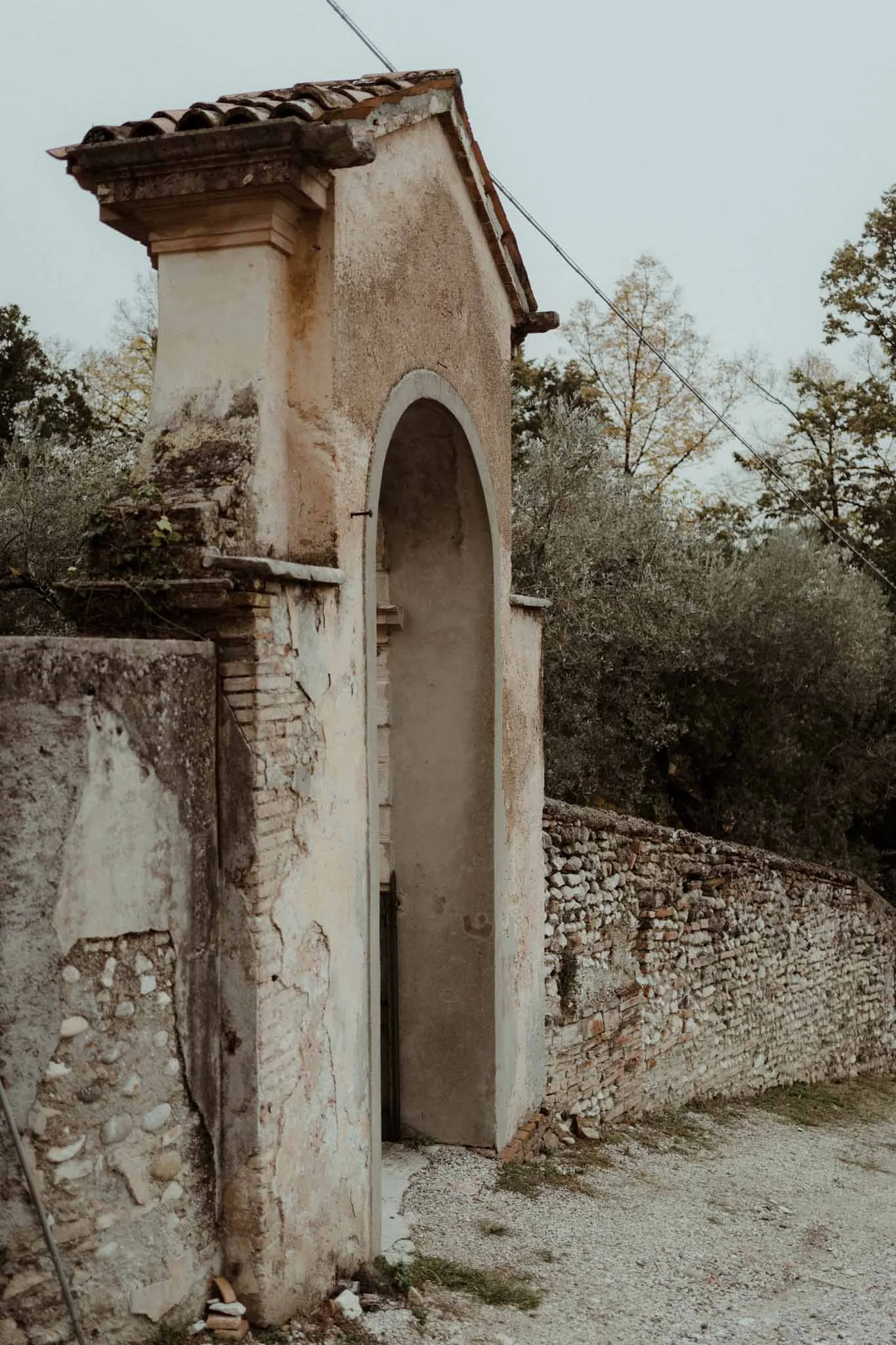 Old, deteriorated stone and stucco archway with tiled roof, part of a crumbling courtyard wall, trees in the background, and a wire running diagonally across the sky.