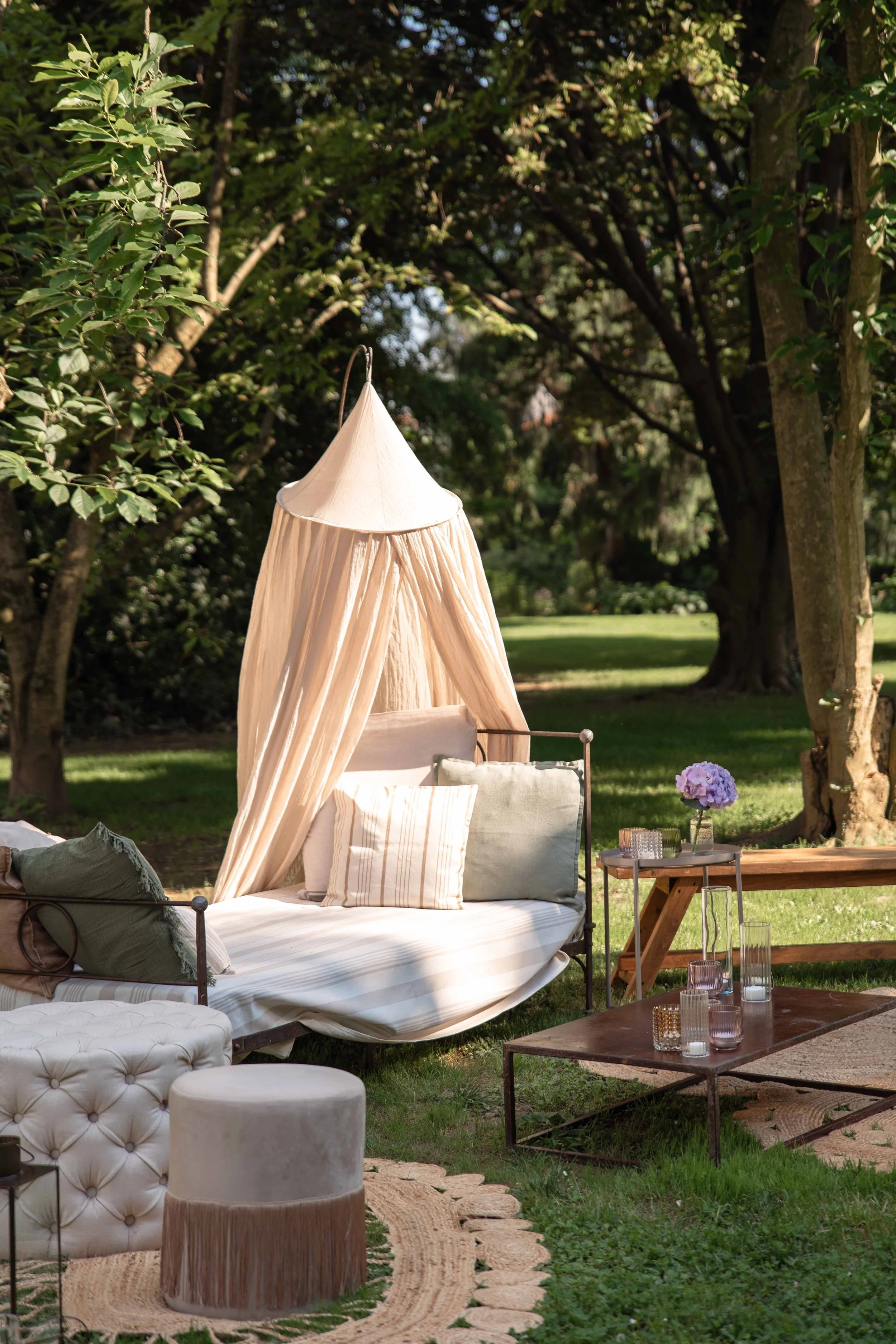 Outdoor seating area in a garden with a canopy tent, cushions, a tufted ottoman, a wooden table with glass jars and a purple flower arrangement, and trees in the background.