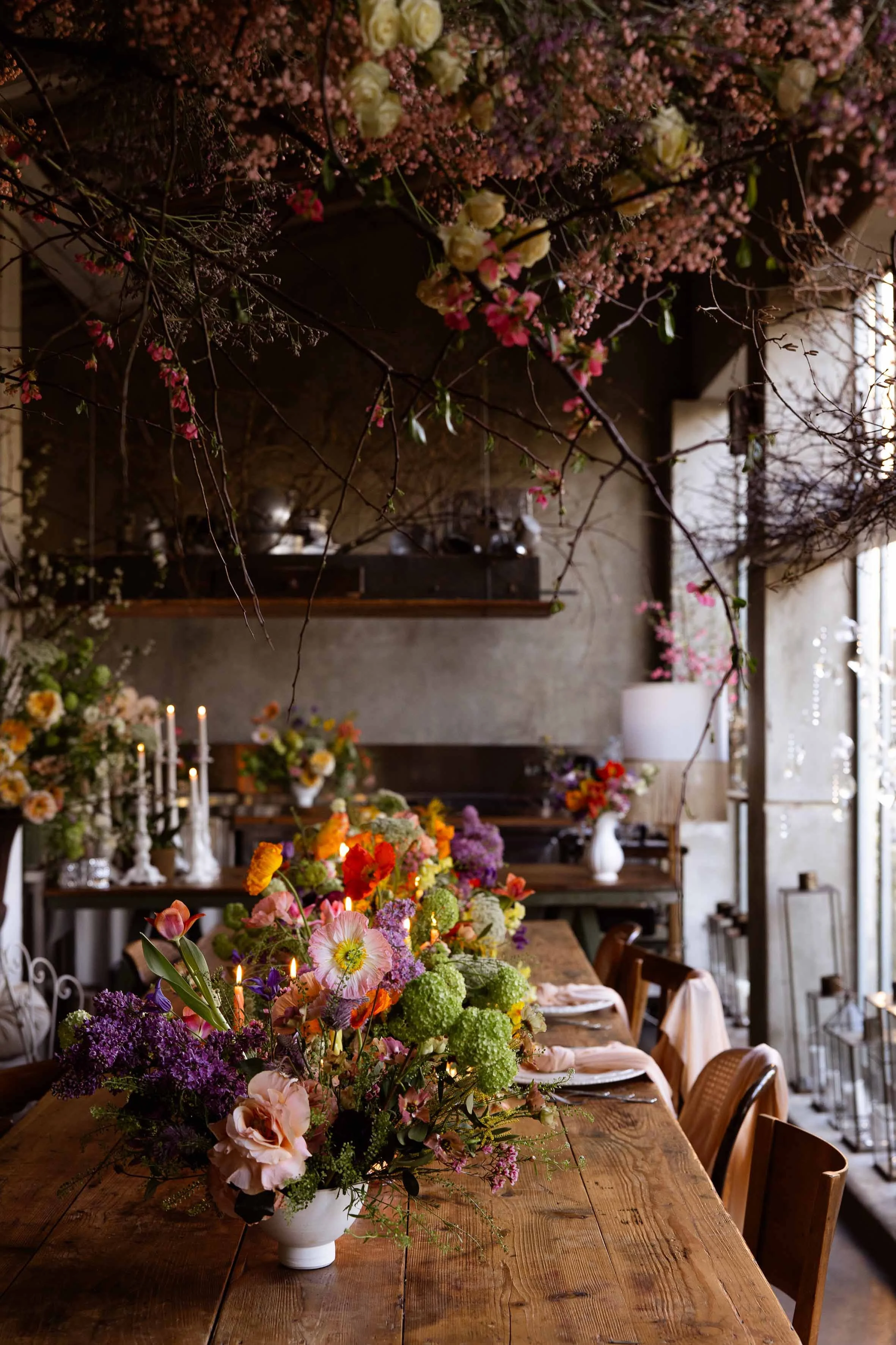 A large floral arrangement on a wooden table in a rustic indoor setting with more floral arrangements and table settings in the background.