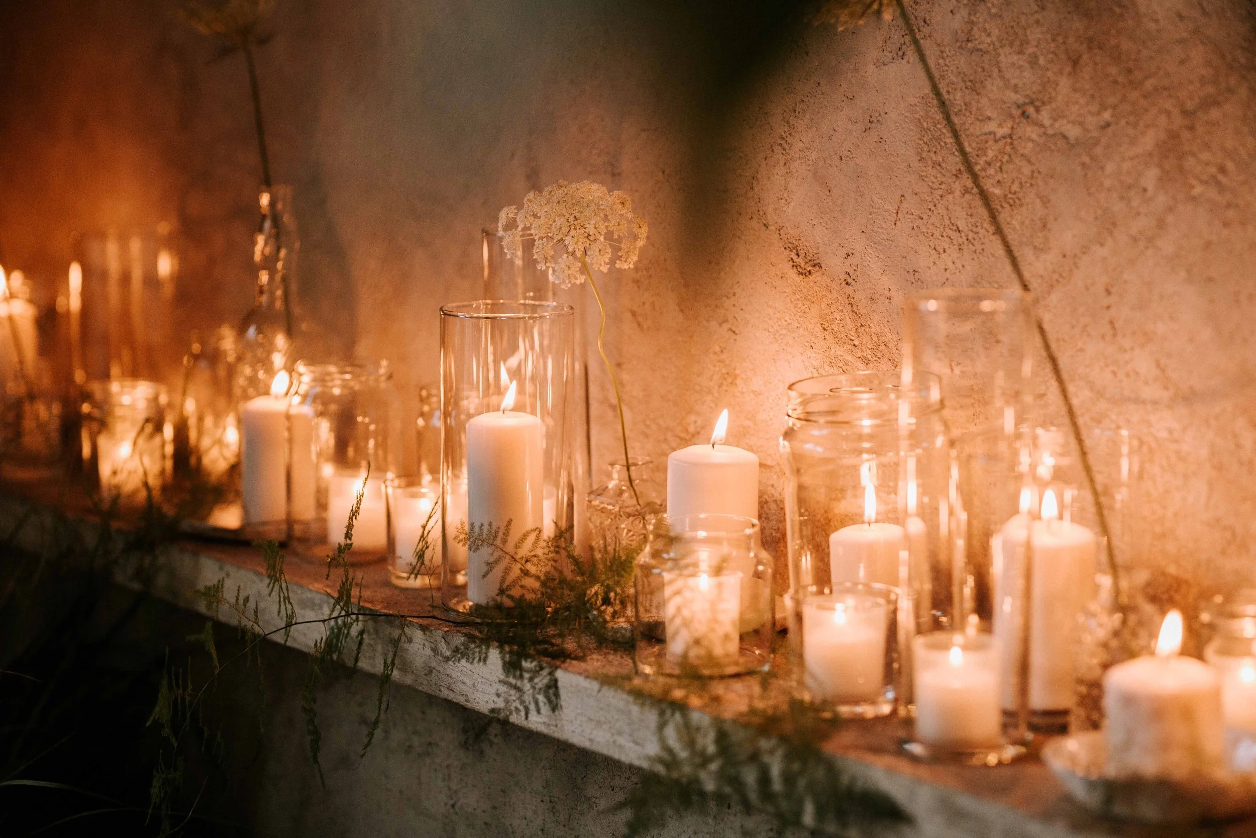 A row of white candles in glass holders and a single white flower in a tall glass vase on a wooden surface against a textured wall, lit softly for ambiance.