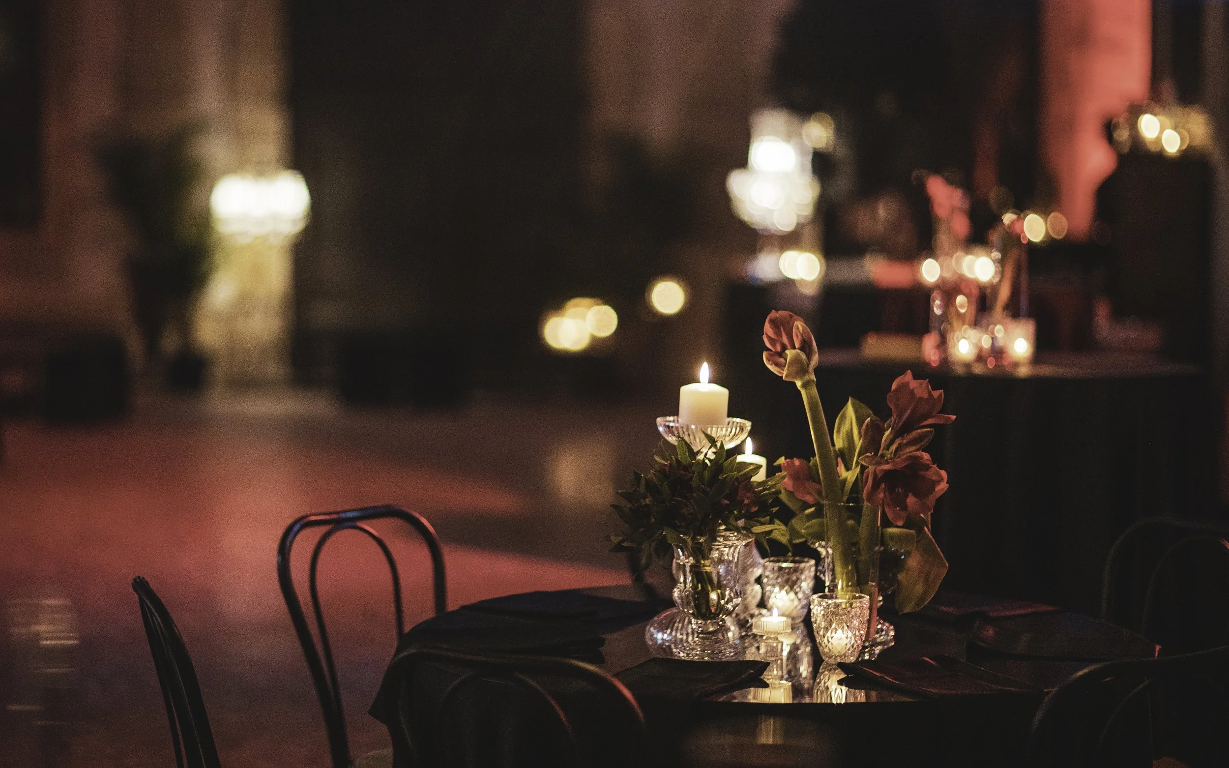 A dimly lit event space with a table decorated with candles, flowers, and glassware. Soft, warm lighting creates a cozy atmosphere.