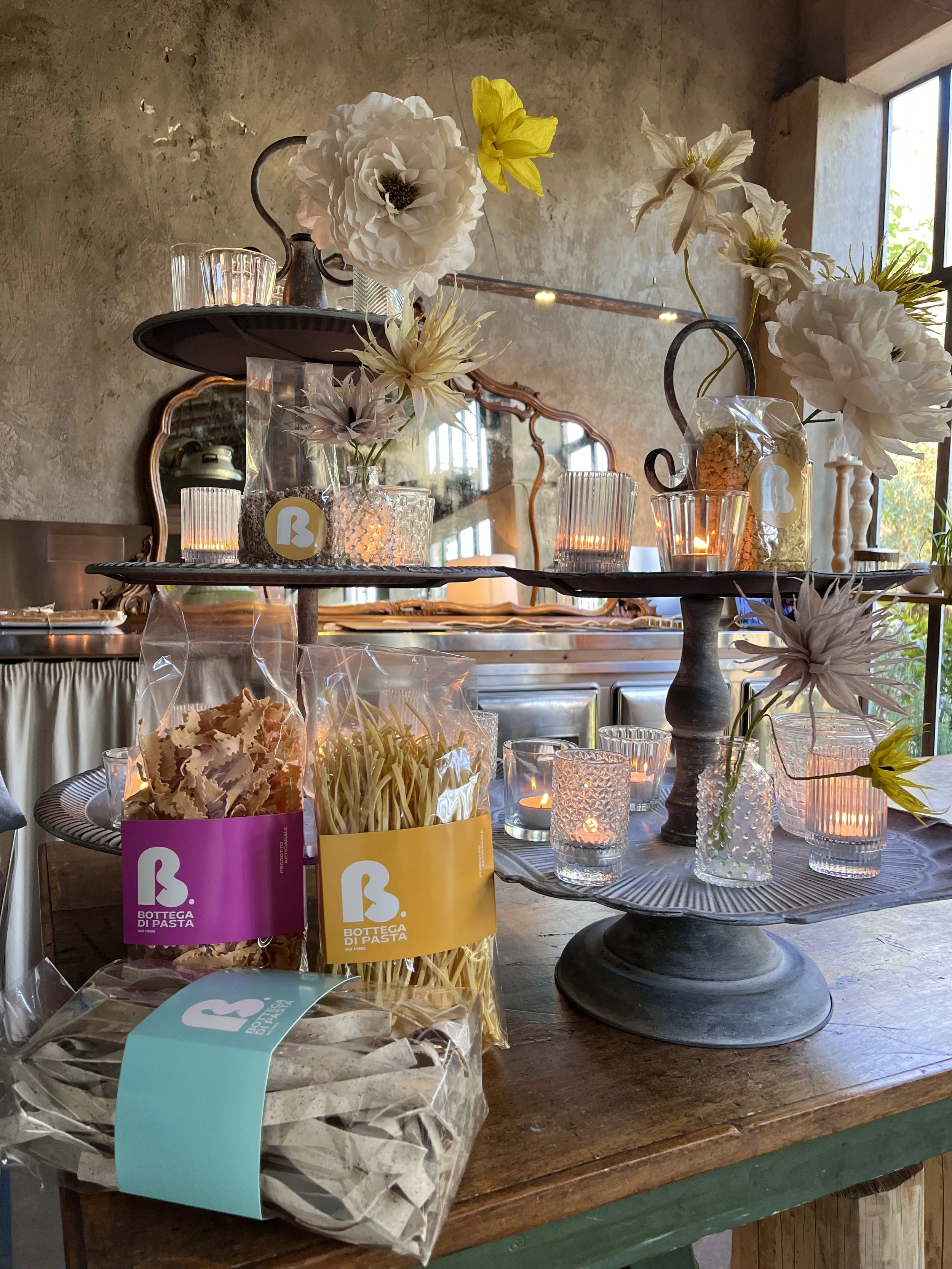 Display of various flower arrangements and containers on a wooden table, with candles and a mirror in the background in a rustic setting.