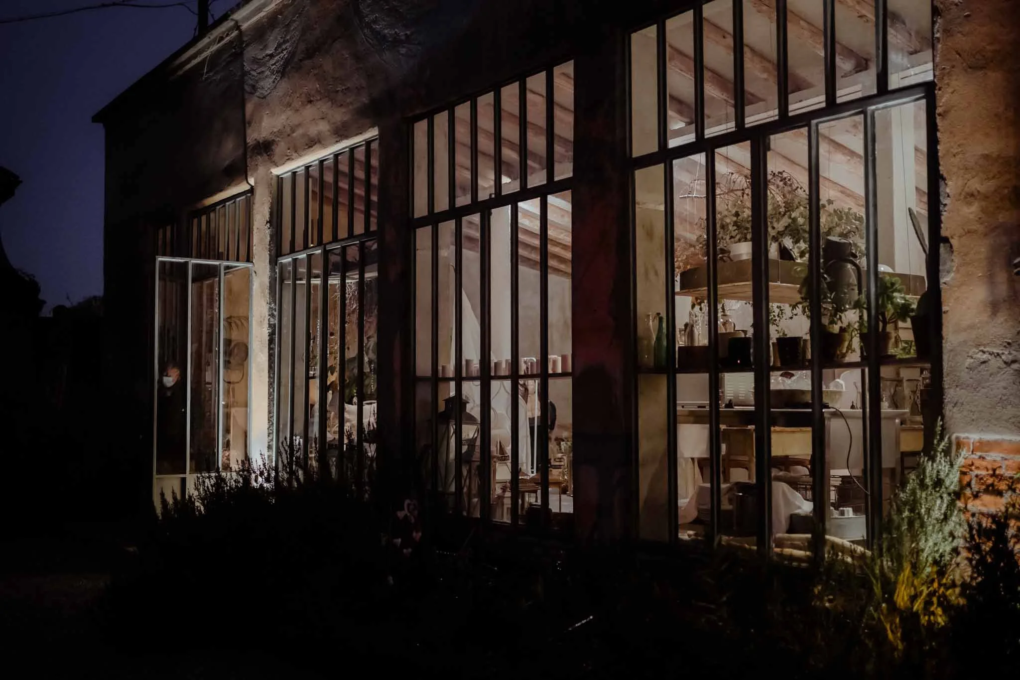 Night view of a house with large glass windows showing an illuminated interior with plants and furniture.