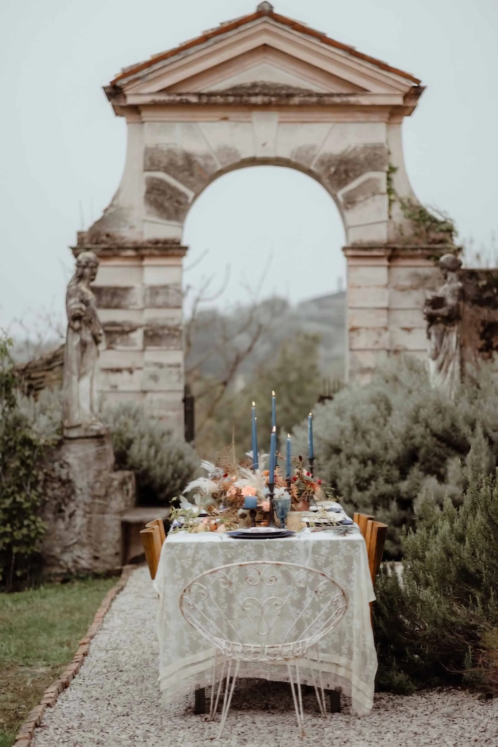 An outdoor dining table set for a meal with candles and floral arrangements, situated along a gravel pathway surrounded by greenery, with an ancient stone arch and ruins in the background.