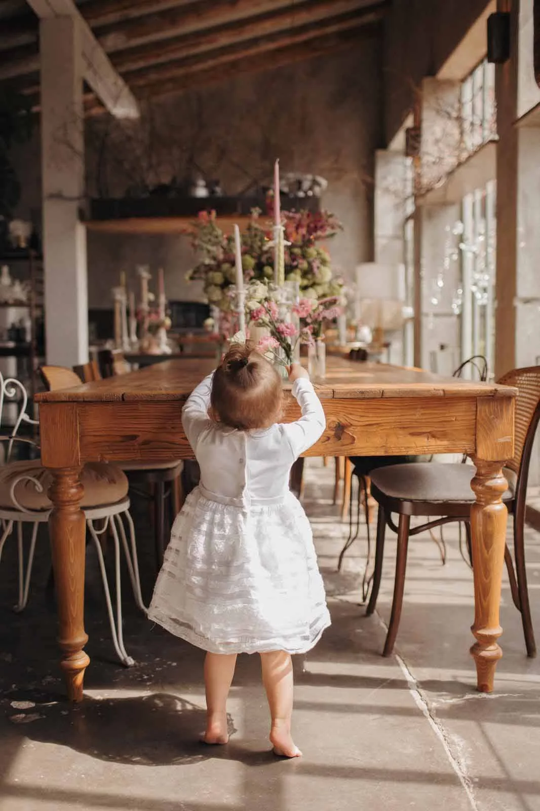 A young girl wearing a white dress, standing barefoot in a rustic dining room, reaching up to adjust a floral centerpiece on a wooden table with pink flowers and candles.