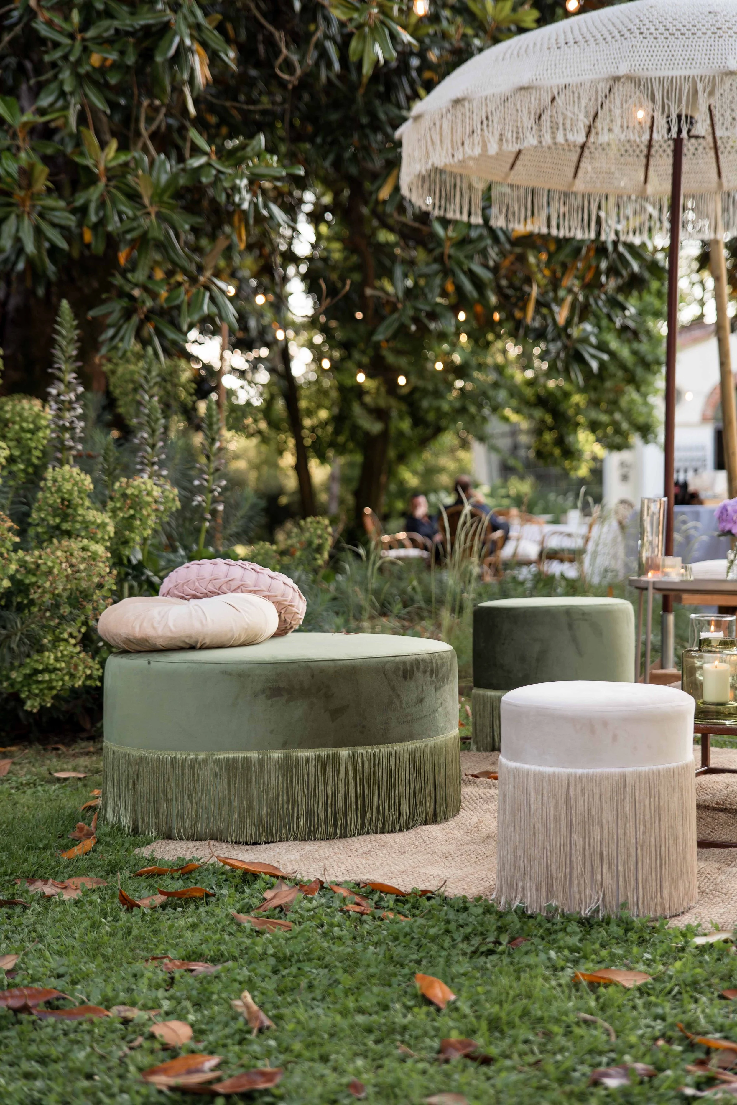 Outdoor patio seating area with velvet ottomans and cushions under a white fringed parasol, surrounded by lush greenery and string lights.