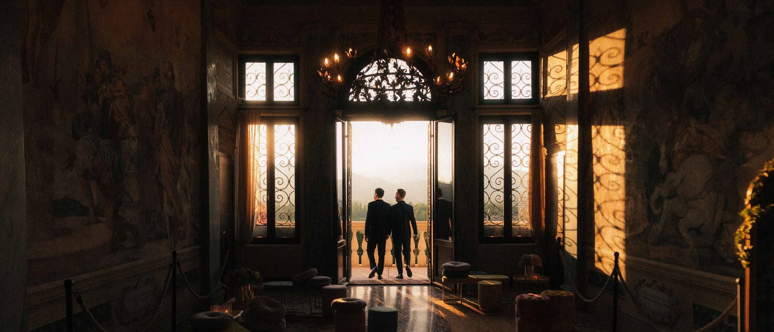 Two men dressed in suits walking through an open door onto a balcony at sunset, with an ornate chandelier hanging from the ceiling and large windows with decorative ironwork.