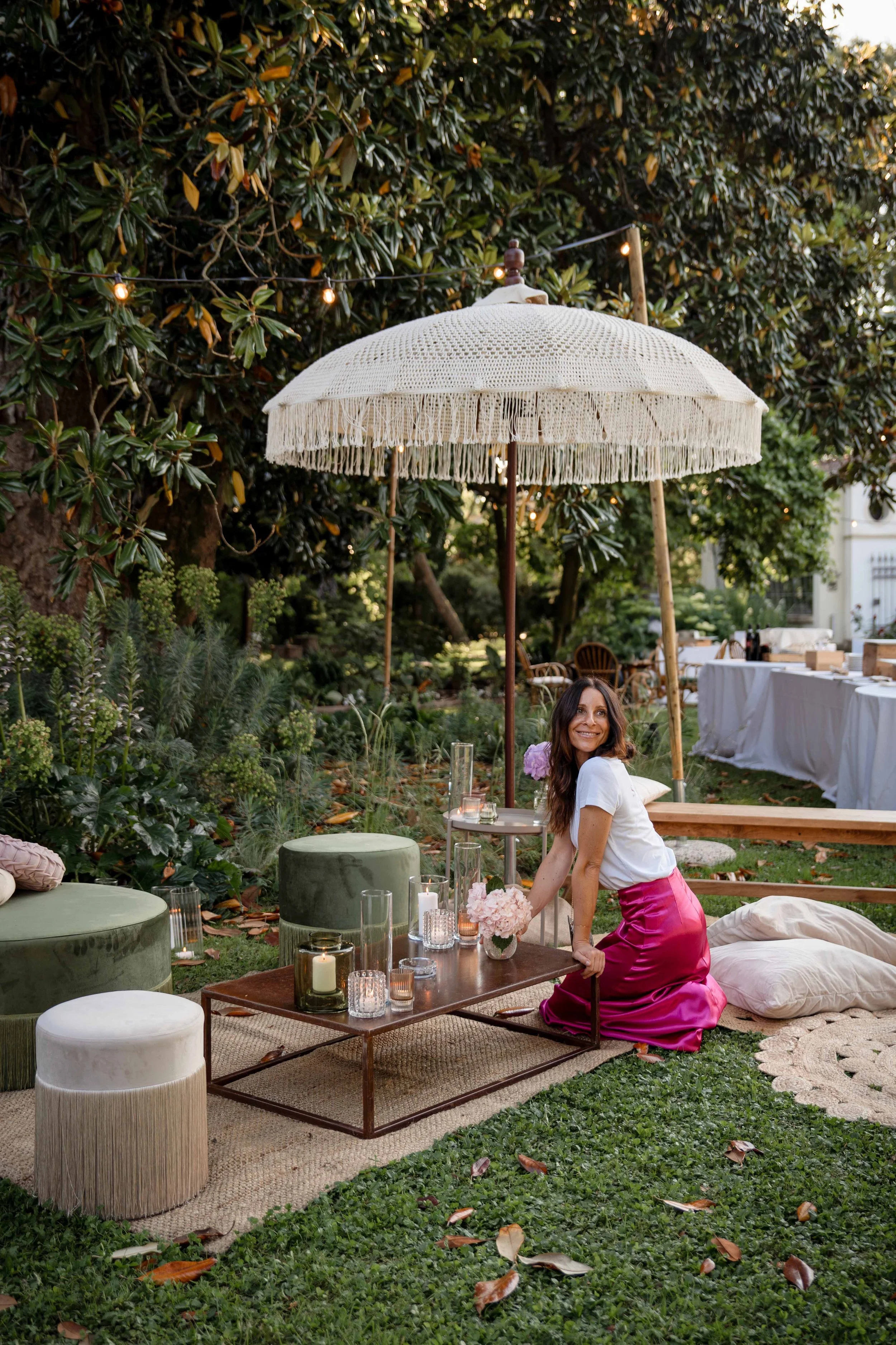 Woman in a white top and pink satin skirt, sitting on the ground at an outdoor garden party with candles, flowers, and a large white umbrella.