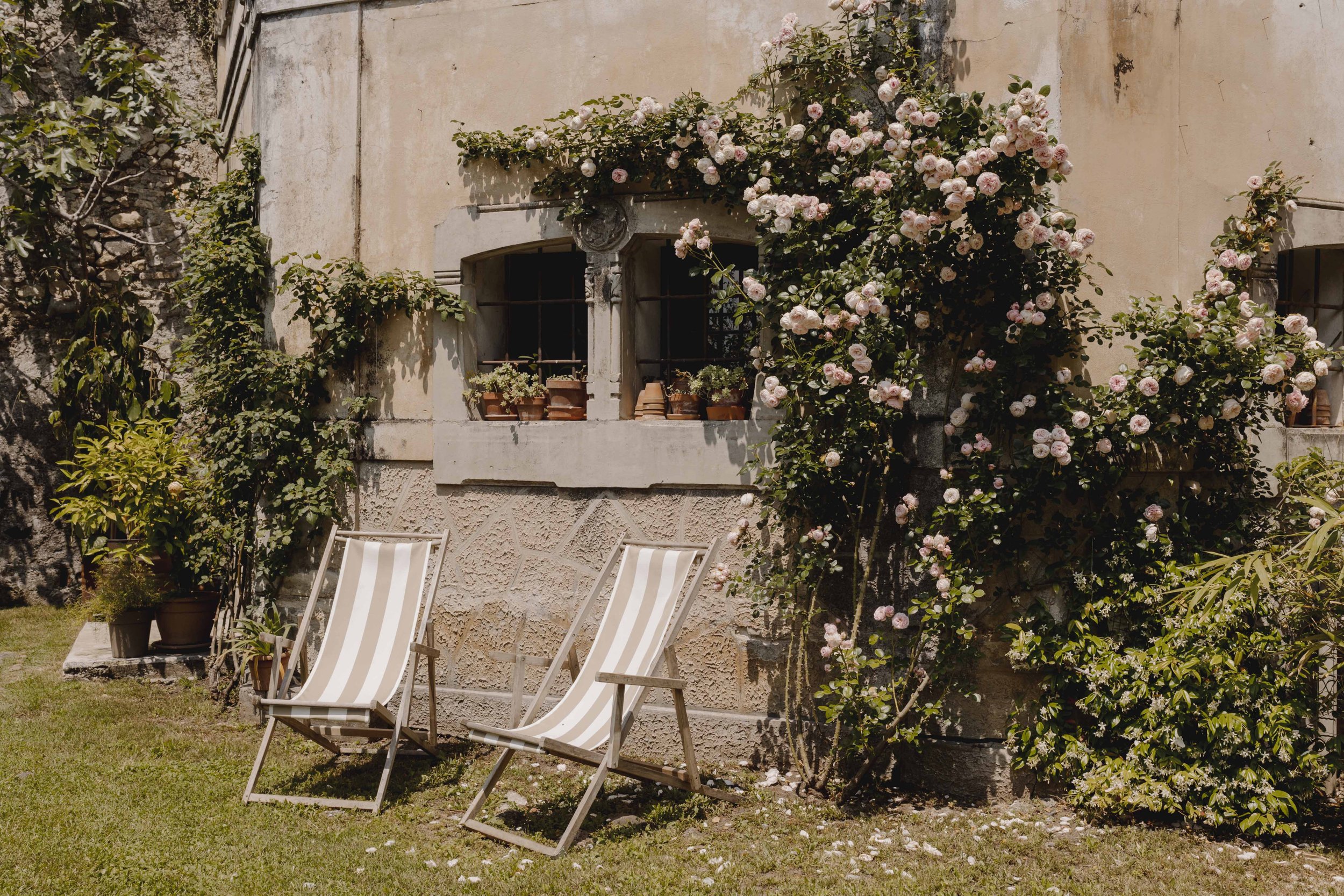 Sunlit outdoor patio featuring two striped deck chairs, potted plants, a vine with pink flowers climbing a textured barn-like wall with windows and flower boxes.