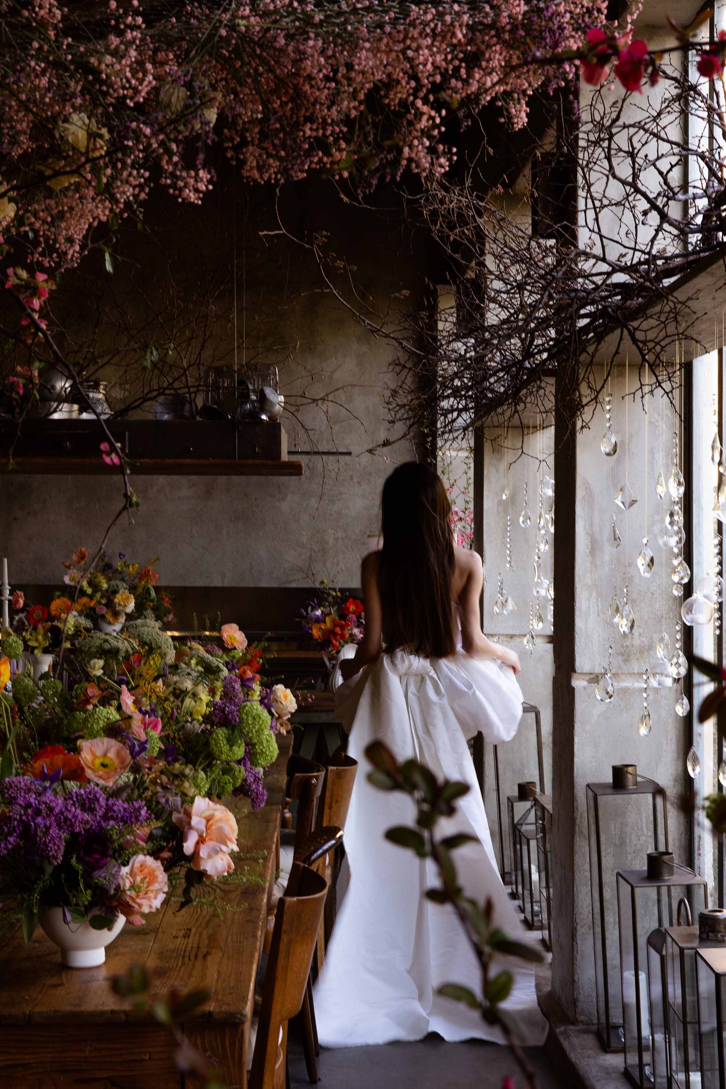 A woman in a white dress sitting in a flower-filled room with hanging crystals, exposed concrete walls, and a large window letting in natural light.
