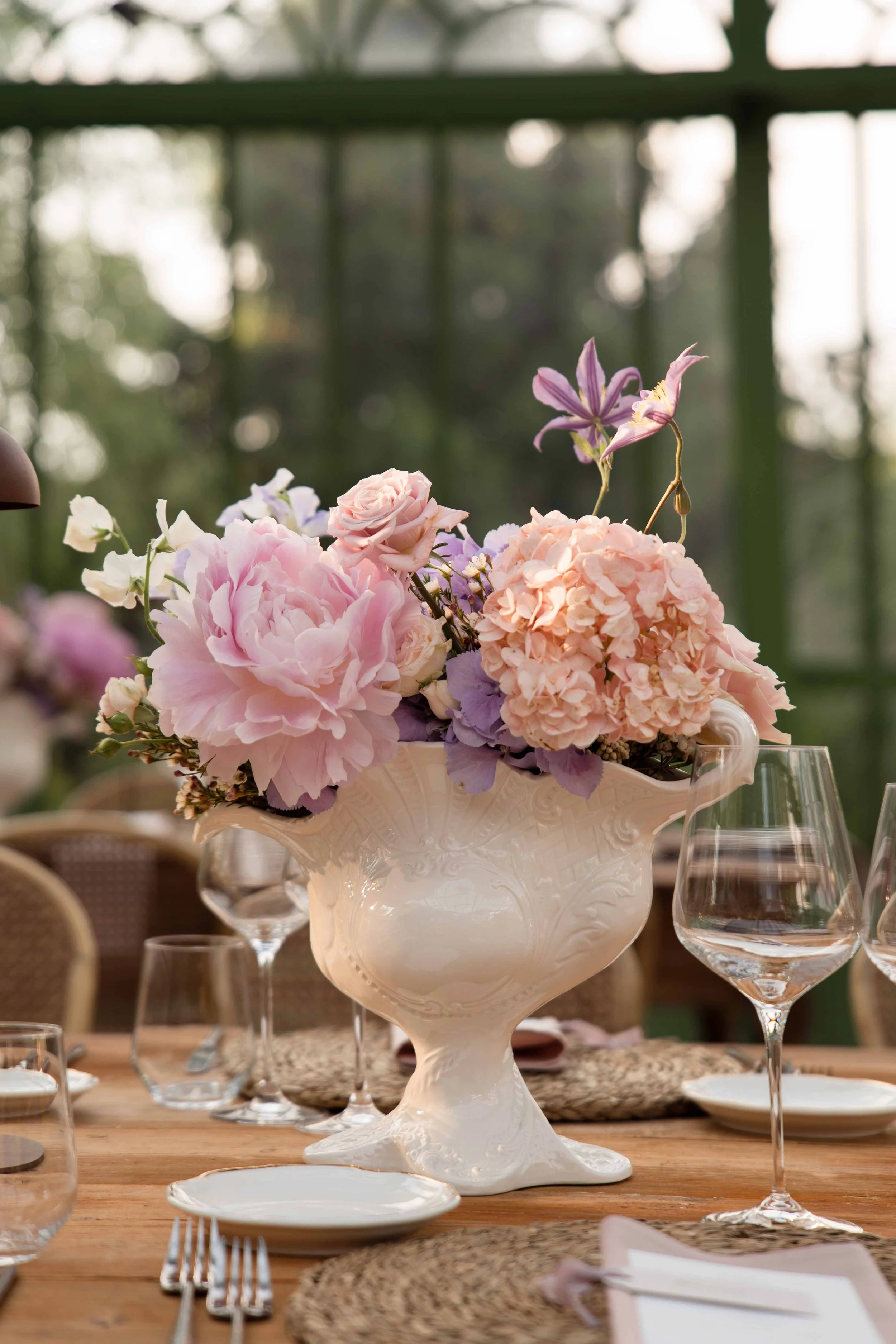A floral centerpiece featuring pink and purple flowers in an ornate white ceramic vase, surrounded by glassware, plates, and cutlery on a wooden dining table set for a meal. A blurred outdoor setting is visible in the background.