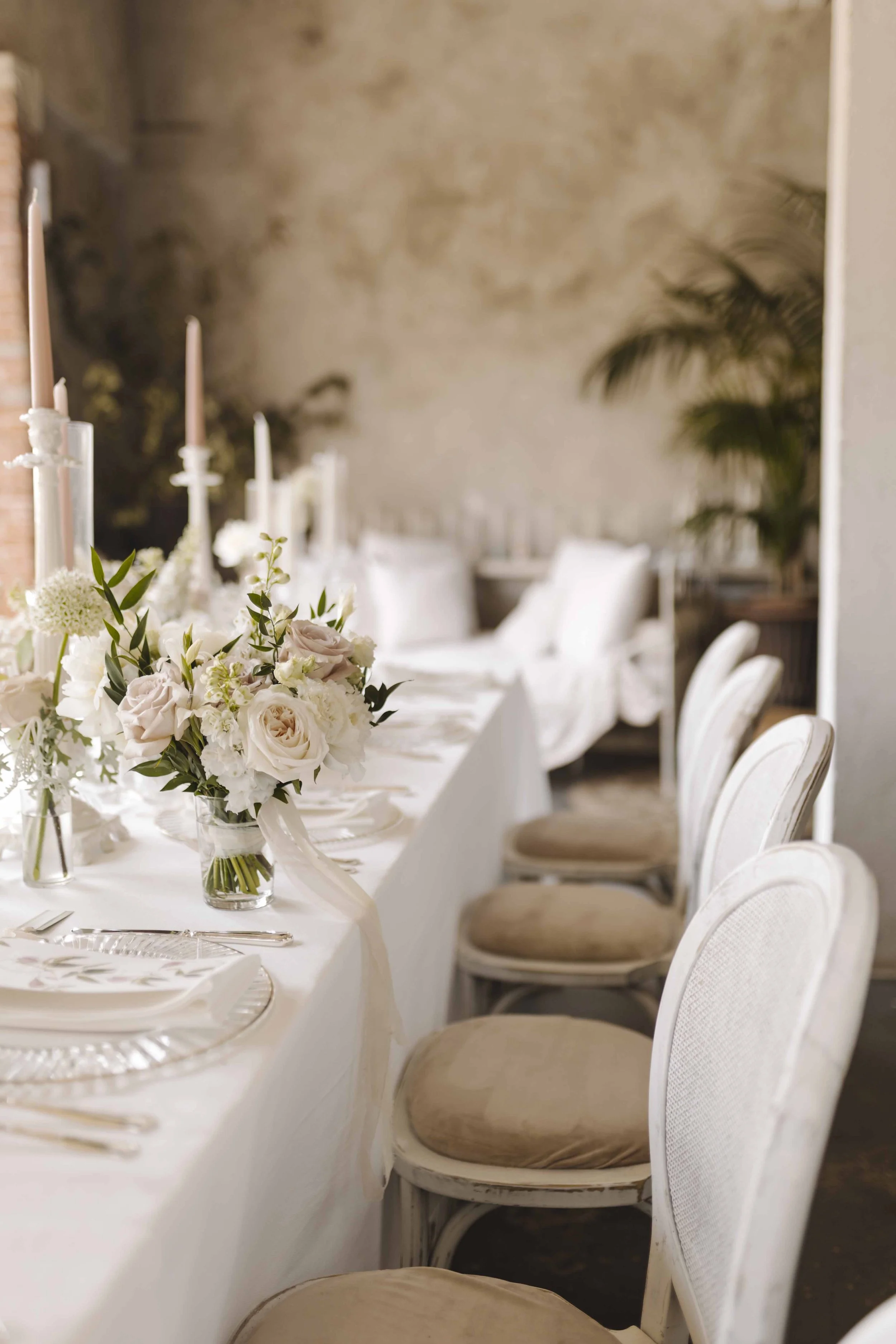 A wedding reception table decorated with a floral centerpiece, candles, and tableware, set in a rustic indoor venue with white chairs and a beige wall with greenery in the background.