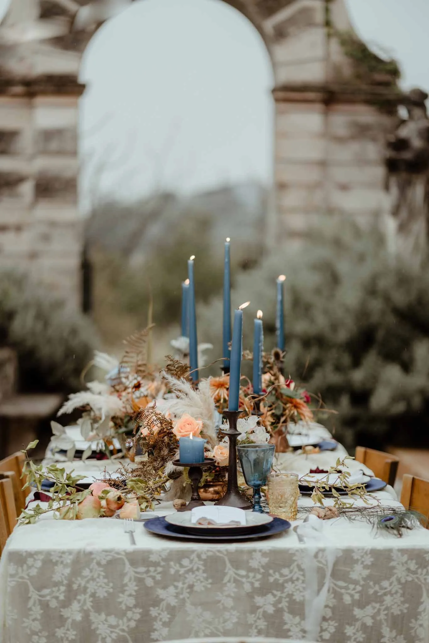 A decorated table set for a celebration with candles, flowers, and tableware, with a background of an arch and ruins.