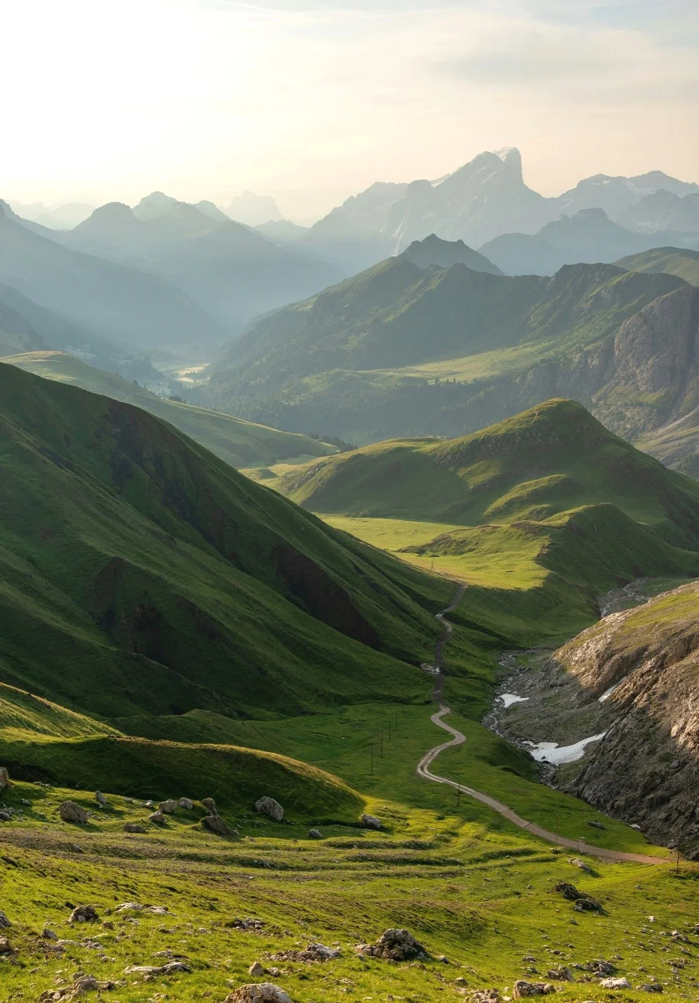 A mountainous landscape with lush green hills, a winding dirt road, and a river flowing through the valley, with distant mountain peaks under a partly cloudy sky.