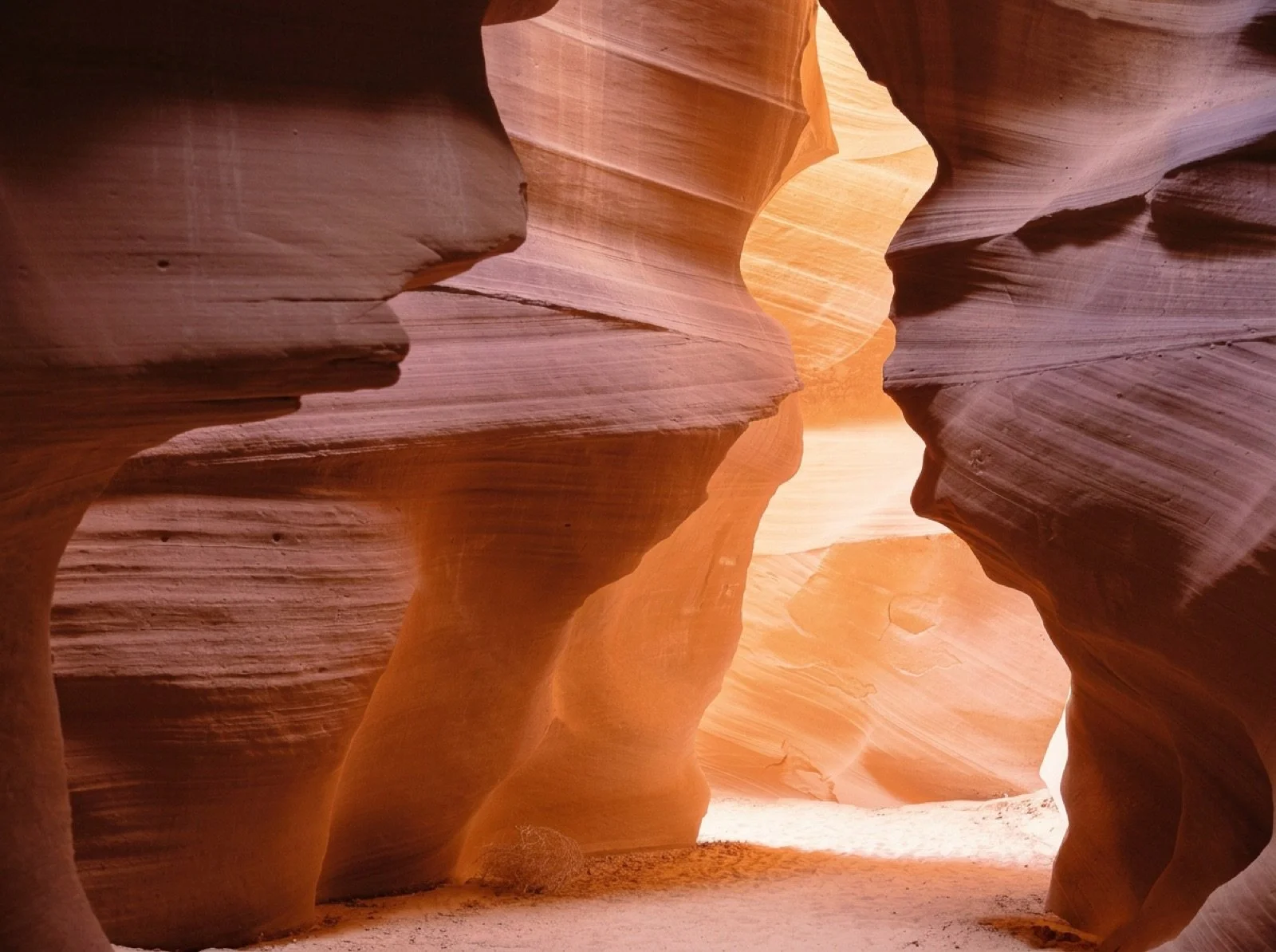 Narrow slot canyon with smooth, layered sandstone walls in shades of orange and red, sunlight filtering through the opening.