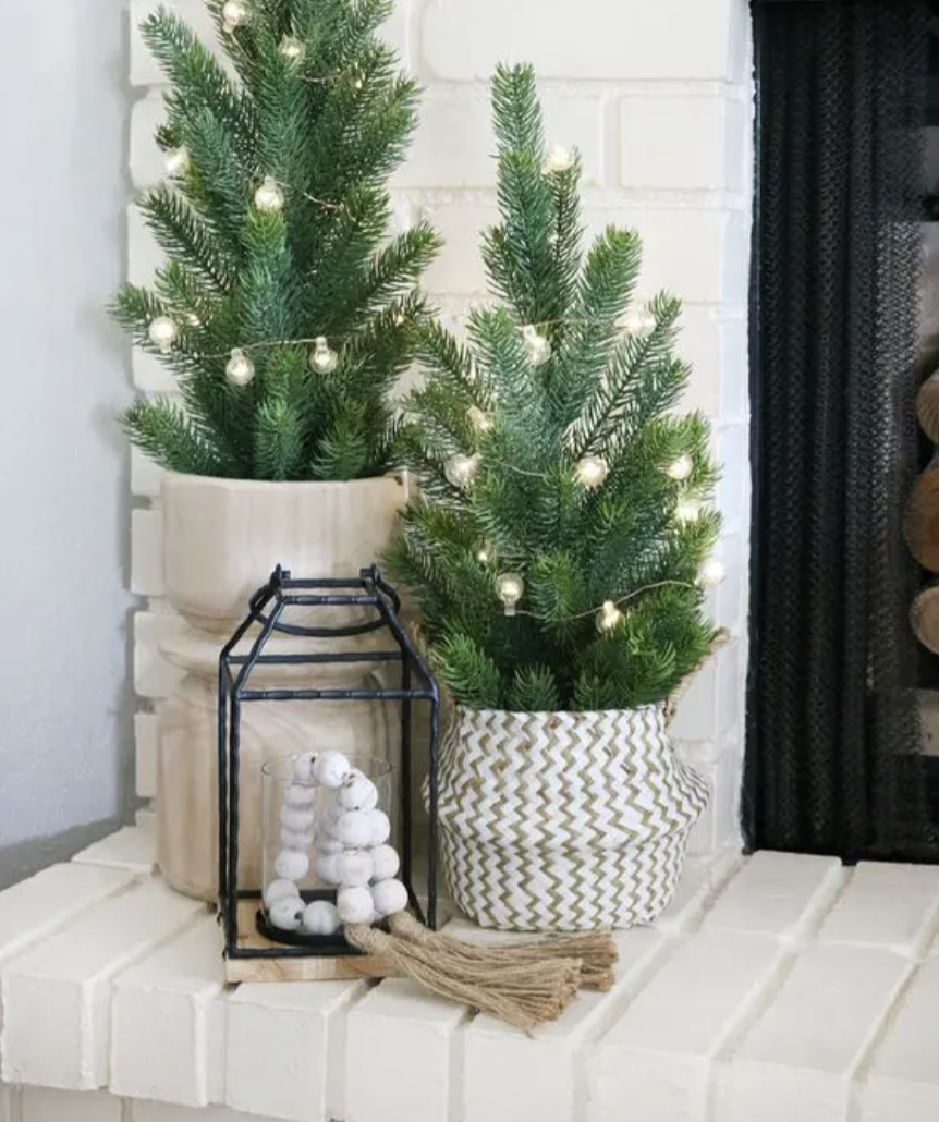 Two small decorated Christmas trees in patterned pots, with a black lantern holding a white garland and a tassel, placed on a white brick surface near a white brick wall.