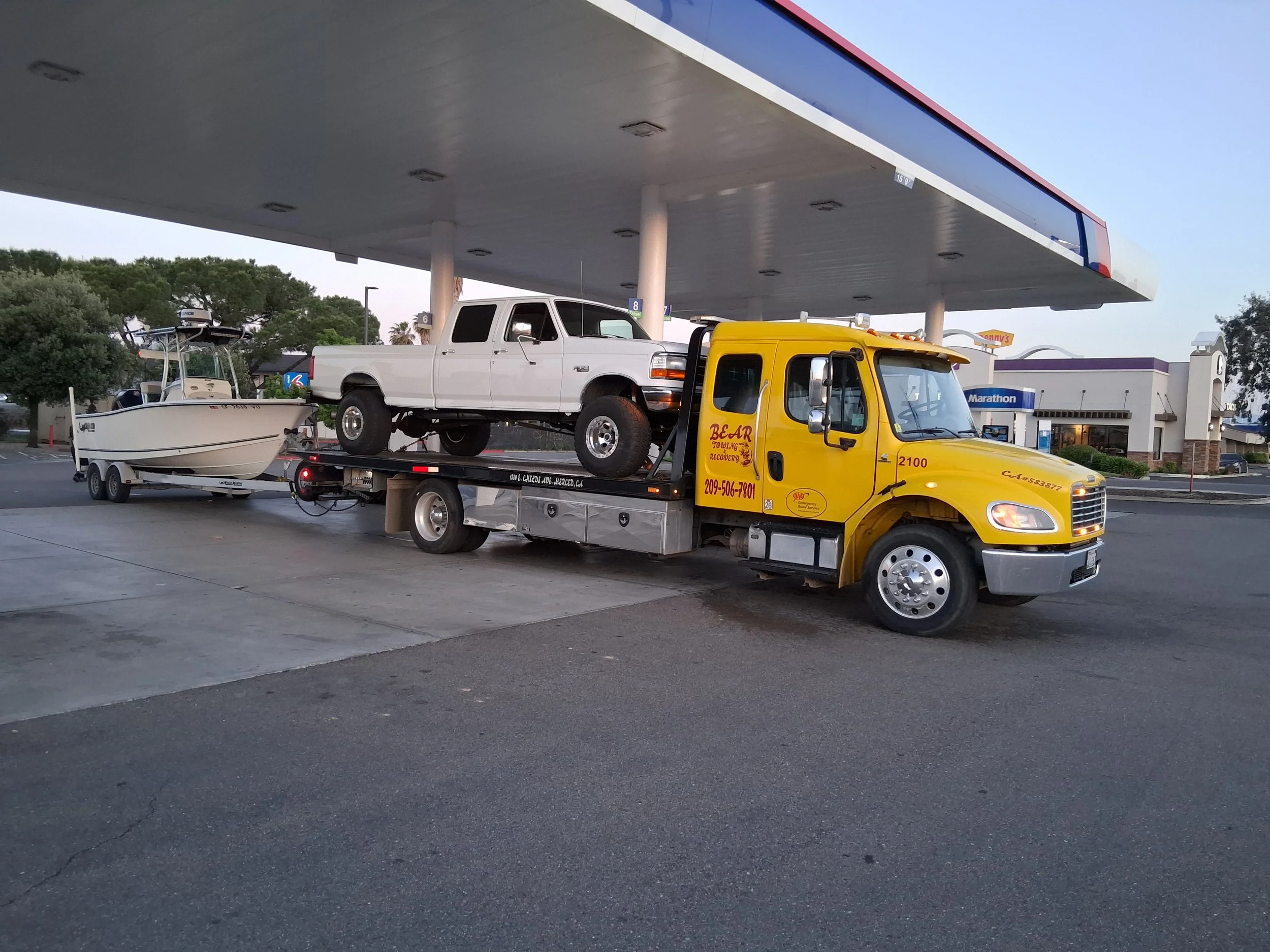 A yellow tow truck is carrying a white Ford pickup truck on its flatbed. The tow truck is parked in front of a gas station with a canopy above the vehicles.