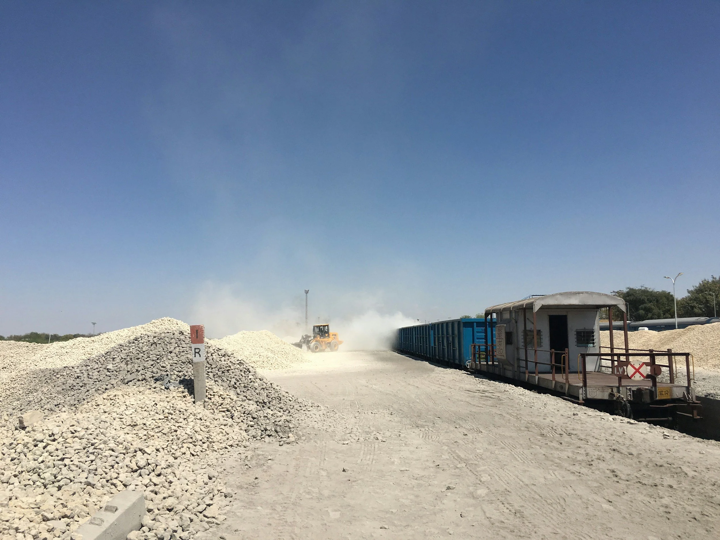 A construction site with piles of gravel and dirt, a forklift, train cars, and a blue sky overhead.
