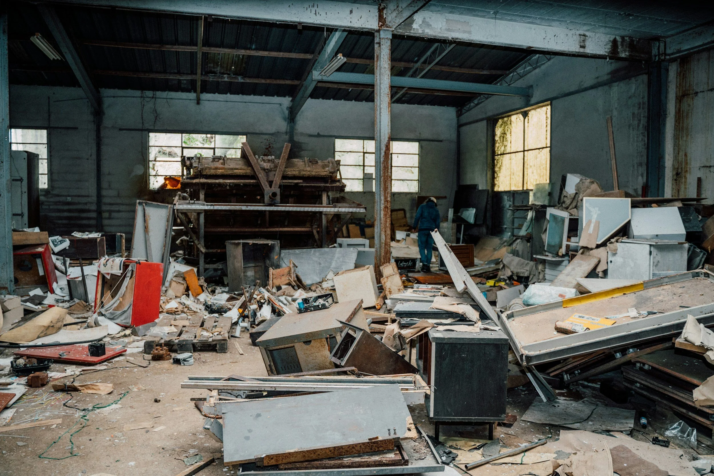 Interior of an abandoned and cluttered building filled with debris, broken furniture, and discarded items, with a person wearing a hoodie and hat walking among the mess.
