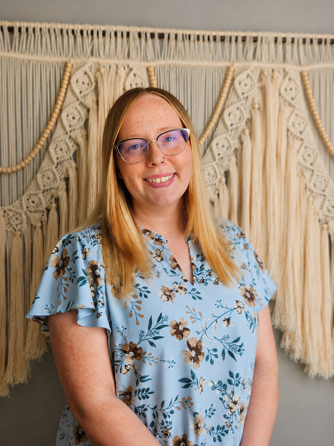 A woman with long red hair and glasses smiling at the camera, standing in front of a large woven macramé wall hanging with beige and cream colors, wearing a light blue floral dress.