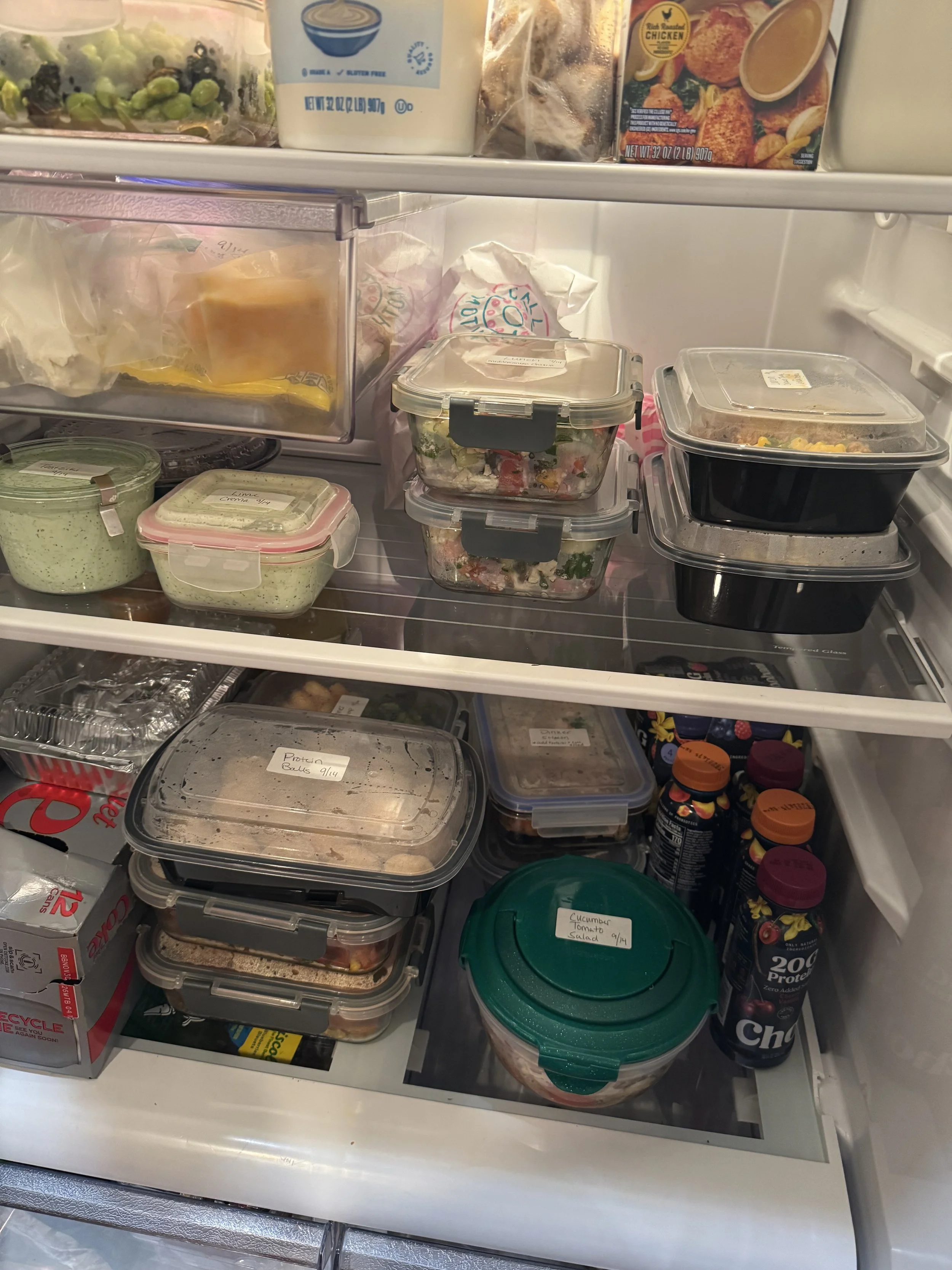 Inside of a refrigerator with various containers of food, including salads, sauces, and leftovers, as well as some bottles of drink and other grocery items.