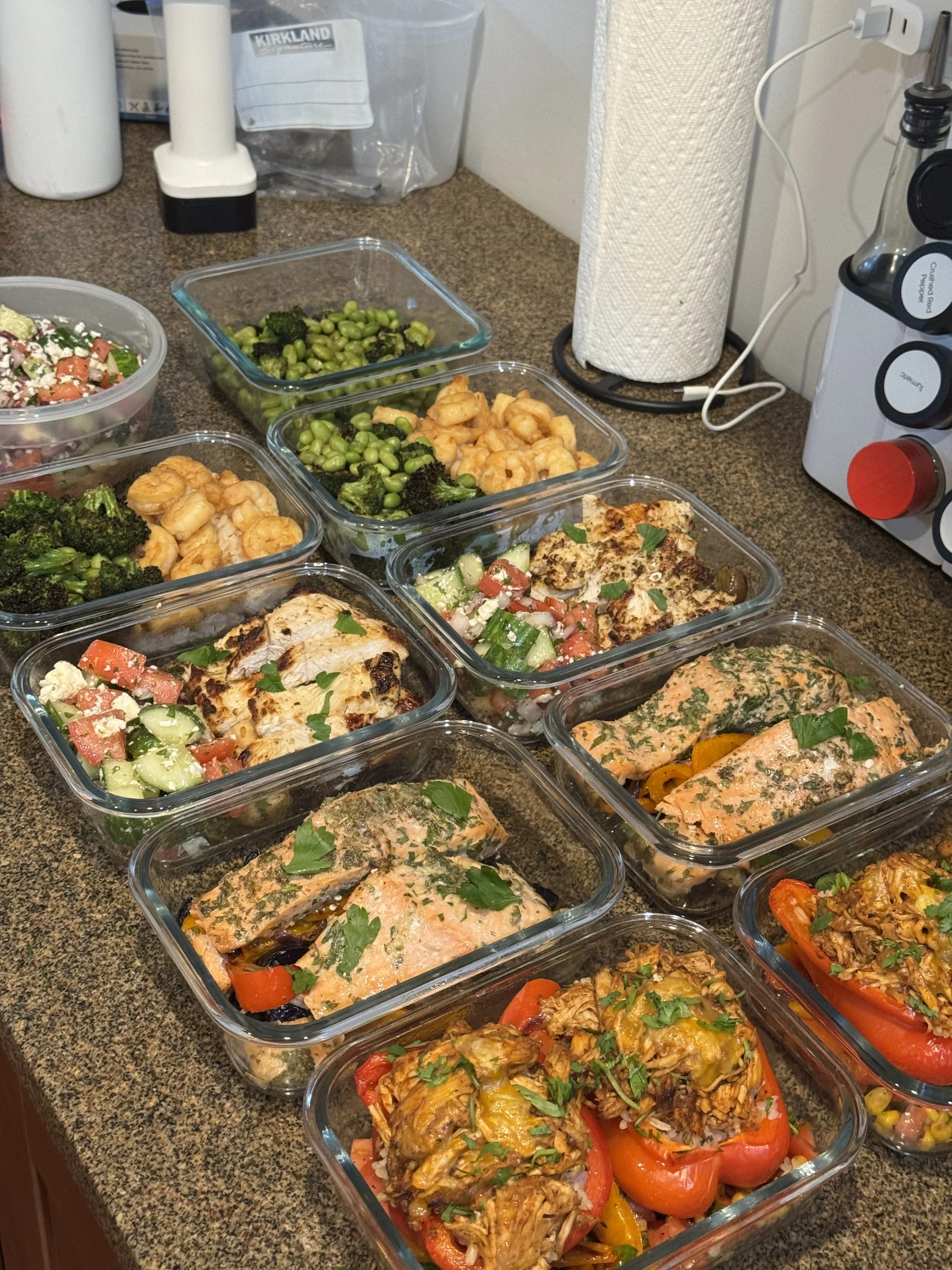 Various cooked dishes arranged in glass containers on a kitchen countertop, including chicken with vegetables, shrimp, broccoli, edamame, and salads.