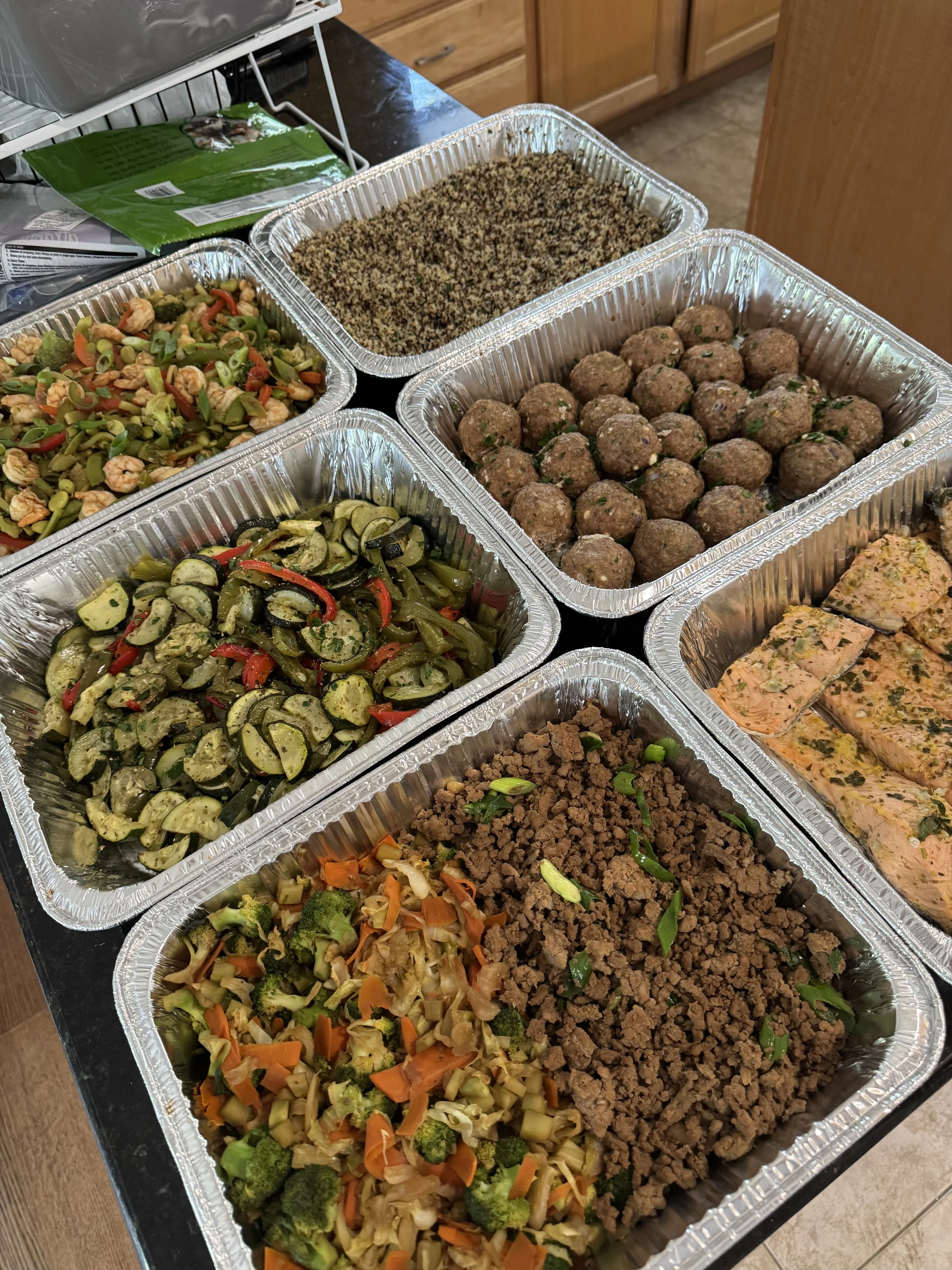 Multiple aluminum foil trays filled with prepared food, including mixed vegetables, meatballs, and cooked ground meat with green onions, arranged on a black countertop.
