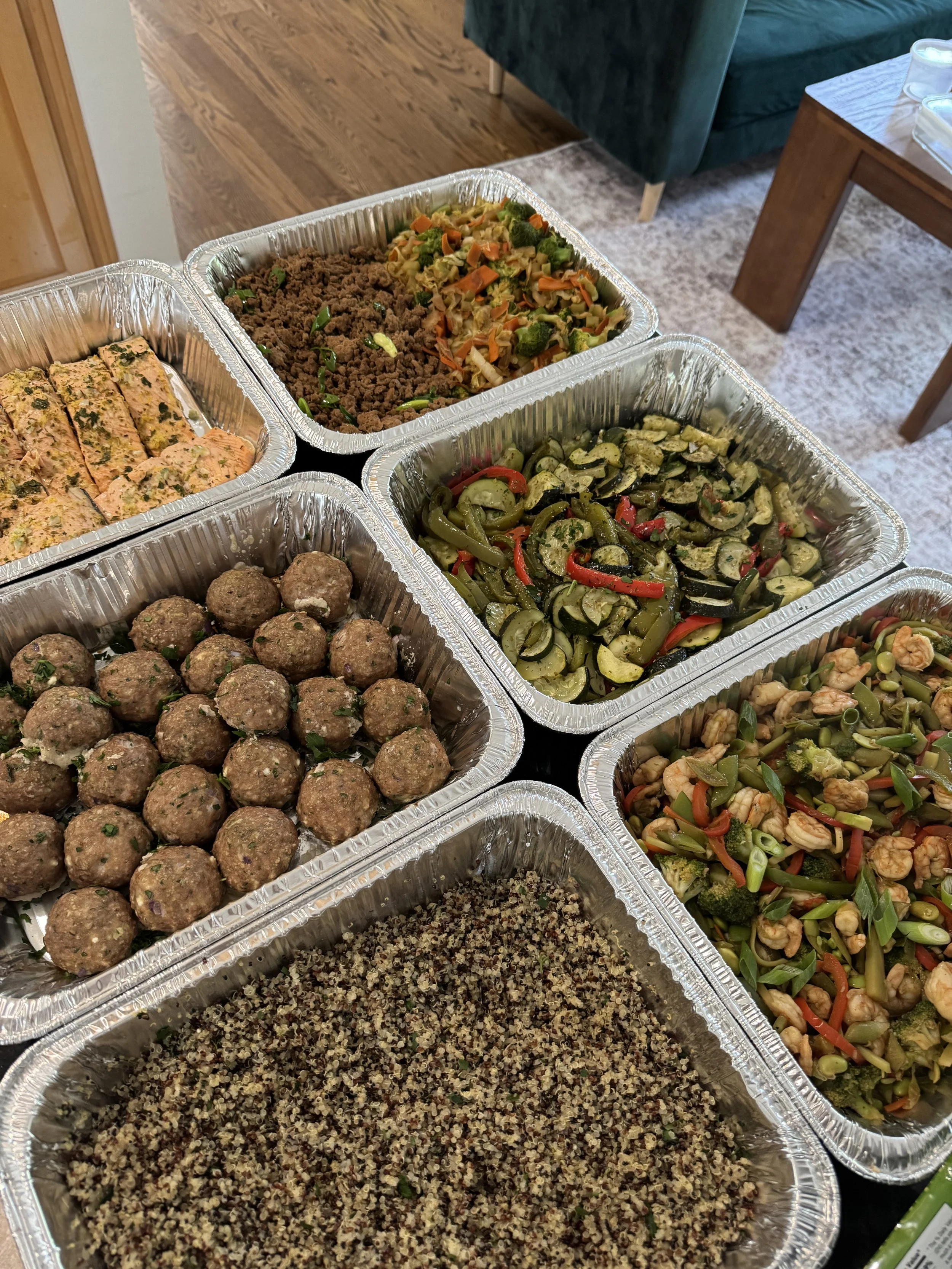 Aluminum trays filled with various cooked foods including grilled vegetables, ground beef with green onions, meatballs, and a quinoa salad, set on a black table in a dining room.