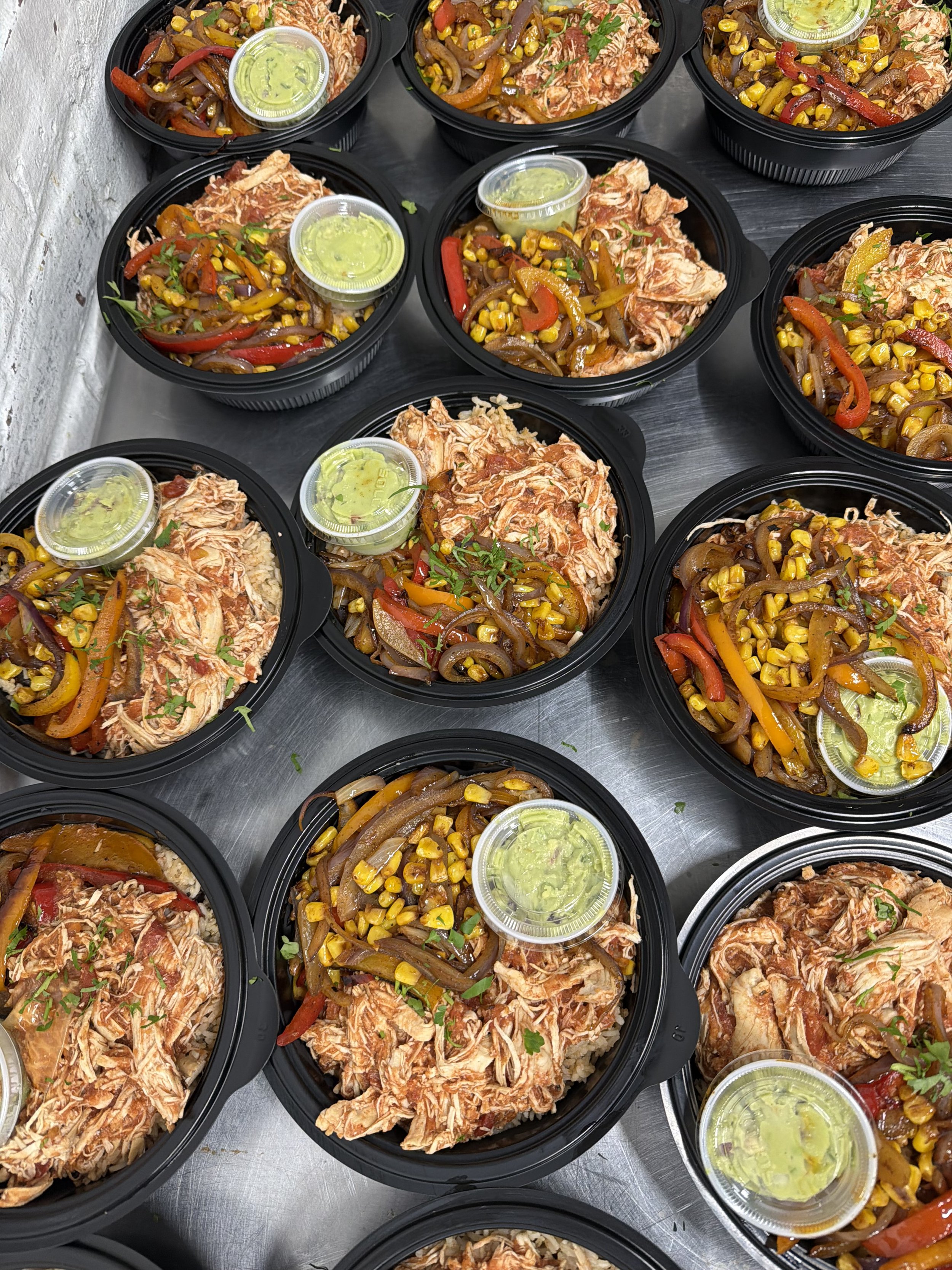 Multiple black plastic food containers filled with shredded chicken, sautéed bell peppers, onions, corn, and a side of green sauce. The containers are on a metallic surface.